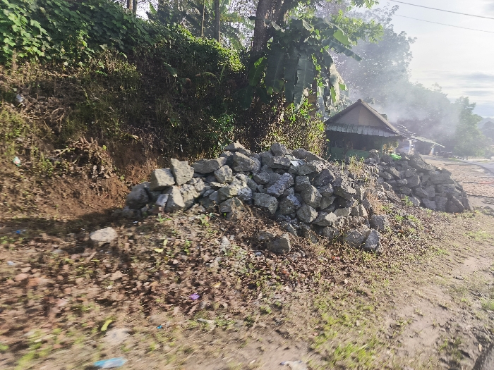The image depicts a pile of gray rocks along the side of a dirt road. The rocks are stacked in a haphazard manner, forming a low wall. The scene is located in the town of Cikalong, Indonesia, at the coordinates -6.767 latitude and 107.442 longitude. The road is covered in dirt and dust, and there is a small amount of green grass growing along the side of the road. There is a small wooden building on the right side of the image, but it is partially obscured by the pile of rocks. The building appears to be abandoned and in a state of disrepair. The surrounding vegetation is thick and green, creating a sense of isolation. There are no people present in the image. The image suggests that the road is rural and not heavily traveled. The pile of rocks could have been used as a retaining wall or to mark a property boundary. The image is taken from a low angle, suggesting the photographer was driving or walking along the road. The image evokes a sense of tranquility and solitude.