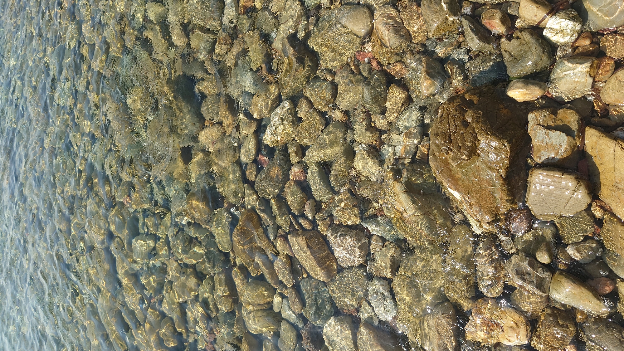 The image shows a shallow body of clear, rippling water flowing over a bed of smooth, multi-colored pebbles and rocks. The water is translucent, allowing a clear view of the stones beneath the surface, some of which are polished and reflect the sunlight. The pebbles vary in size, shape, and color, ranging from light beige and grey to darker brown and reddish hues. The texture of the stones is rough and uneven, with some showing distinct stratification. The overall scene suggests a natural watercourse or a shallow coastal area. The location is identified as Biddeputzi/Villaputzu, Italy, which is a municipality on the island of Sardinia. The image captures a moment of natural beauty with no human presence or activity.