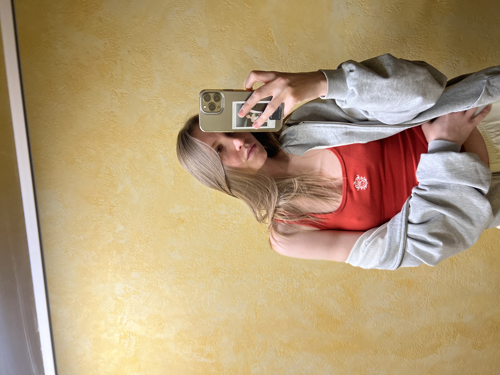 A young, light-skinned woman with blonde hair is taking a selfie. She is wearing a red tank top and a grey zip-up jacket. Her phone is held up, obscuring part of her face, and has a polaroid-style photo attached to the back. The background consists of a textured, yellow-toned wall and a white door frame on the left. The location is not identifiable as Khimki, Russia.