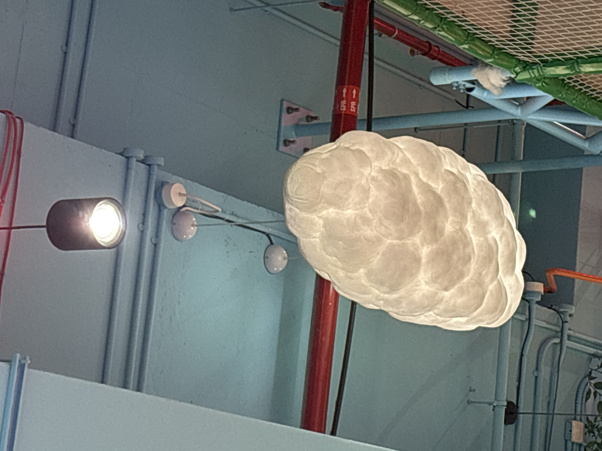 This is a close-up, vertical, eye-level shot of a cloud-shaped light fixture hanging from a light blue ceiling. The fixture is white and appears to be made of a soft, textured material, giving it a realistic cloud-like appearance. Above and behind the cloud fixture, a dark gray cylindrical pendant light is visible, also hanging from the ceiling. Several white circular objects, possibly smoke detectors or small lights, are mounted on the ceiling as well. Red pipes run horizontally across the background, and various blue and gray pipes and structural elements are also present. The scene appears to be indoors, possibly in a recreational facility or a themed environment, given the playful nature of the lighting. There is no indication of the city of Dubai, United Arab Emirates.