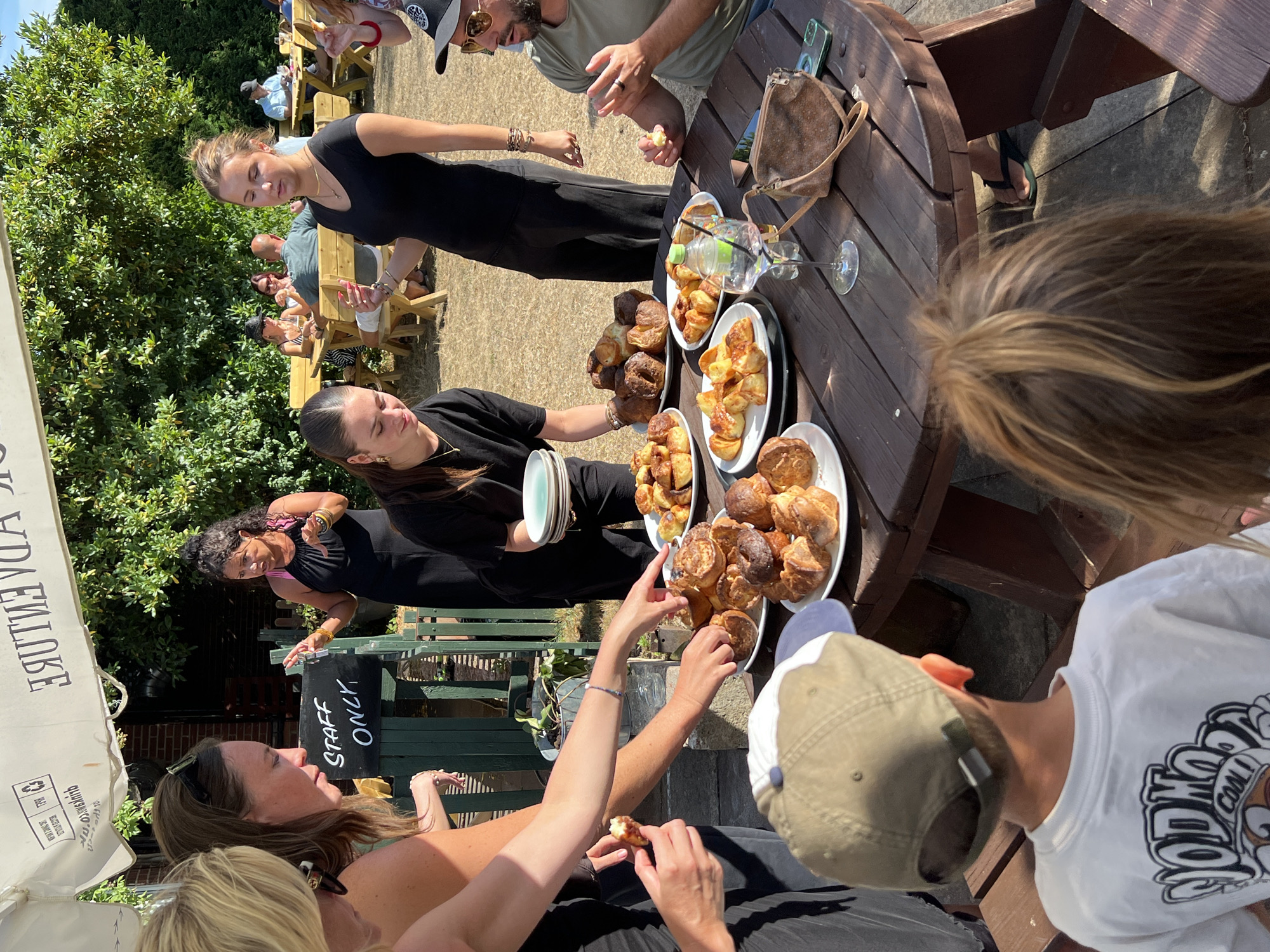 The photograph shows a group of people gathered around a wooden picnic table outdoors, seemingly at an event or a café in Guildford, United Kingdom. The setting appears to be a park or garden area, with lush green trees and bushes in the background. The table is laden with plates of pastries and baked goods, suggesting a catering or buffet setup.
In the foreground, a young boy wearing a white t-shirt with a graphic print on the back and a green baseball cap is seated, facing away from the camera. To his right, the back of another child's head with blonde hair is visible.
On the left side of the table, several adults are interacting. A woman with blonde hair is partially visible, and next to her, a woman with long brown hair and sunglasses on her head is looking down at the table. A woman in a black dress is also present, holding a plate and gesturing with her hands. A woman with dark curly hair, wearing a black sleeveless dress and a pink bra, is standing and speaking, with her hands raised as if explaining something. She appears to be directing or serving the food.
On the right side of the table, a man wearing a green t-shirt and sunglasses is seated, holding a piece of food and appearing to talk. Behind him, another woman in a black dress is visible, and further in the background, other patrons are seated at picnic tables.
The table itself is made of dark wood and has a weathered appearance. On it, there are several plates filled with a variety of pastries, some appearing to be croissants and others like mini Yorkshire puddings or sweet buns. A glass of pinkish liquid with ice, a bottle of water, a small brown purse, and a mobile phone are also on the table. A sign behind the people on the left reads "STAFF ONLY."
The overall lighting suggests it is a sunny day, casting distinct shadows. The atmosphere appears casual and convivial.