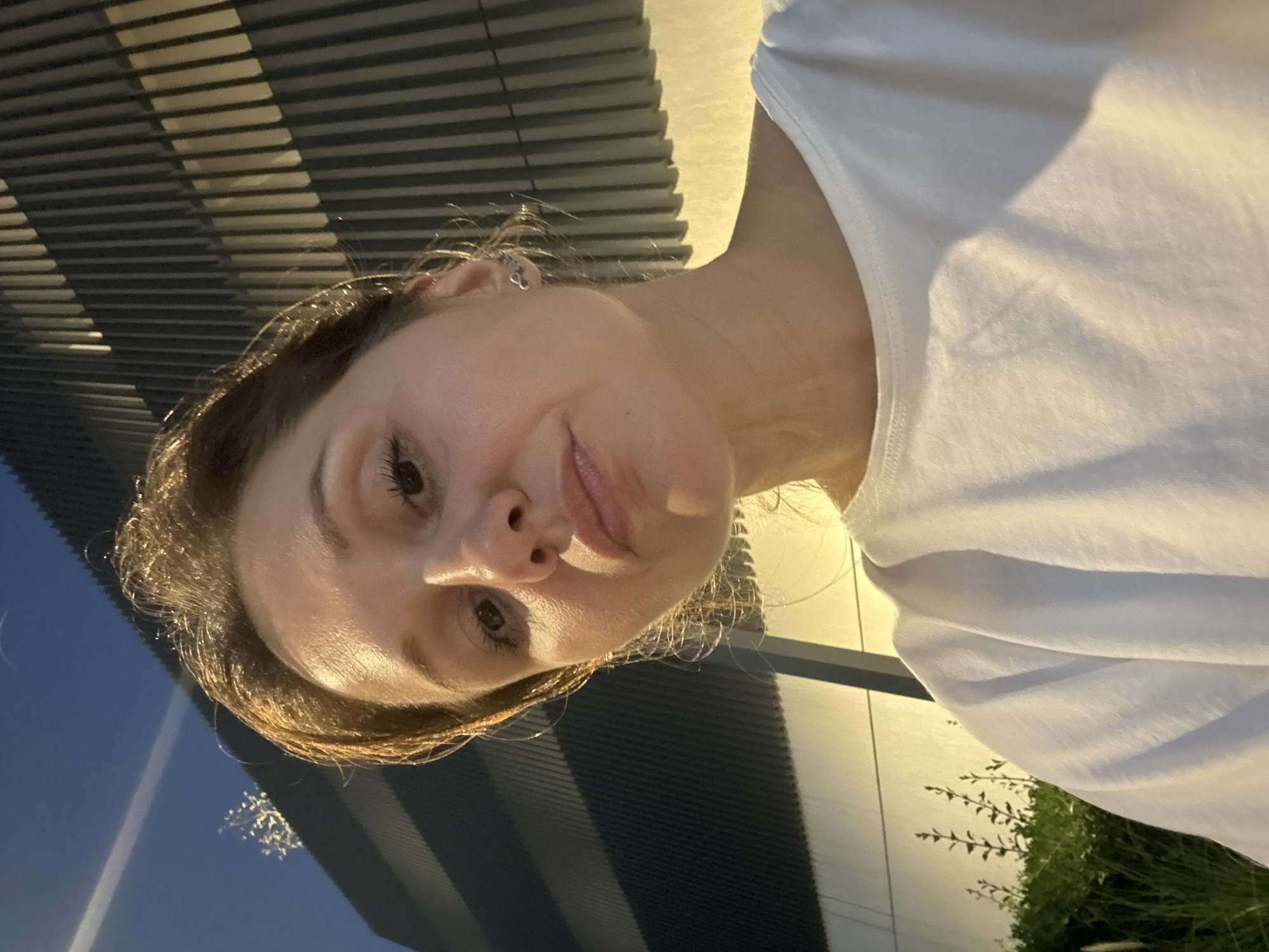 A tilted, close-up portrait features a young woman outdoors in an evening or nighttime setting. The woman, positioned toward the bottom left of the frame, has brown hair and is wearing a white t-shirt. She is looking directly at the camera with a subtle pout or thoughtful expression. A small, silver hoop earring is visible on her left ear.
Warm, artificial light illuminates her face and parts of her hair and t-shirt, creating highlights on her skin, suggesting either the golden hour or early evening under artificial light sources.
The background consists of modern architectural elements. A dark, slatted structure with repetitive horizontal lines occupies the upper and left portions of the image. To the right, a light-colored building facade is visible, also illuminated by warm light. Below this facade, some dark green foliage, possibly bushes or plants, can be seen. The sky, visible in the upper left corner, appears dark blue, consistent with dusk or night. The overall scene suggests a casual selfie taken in an urban environment in Dubai, United Arab Emirates. No text is visible in the image.
