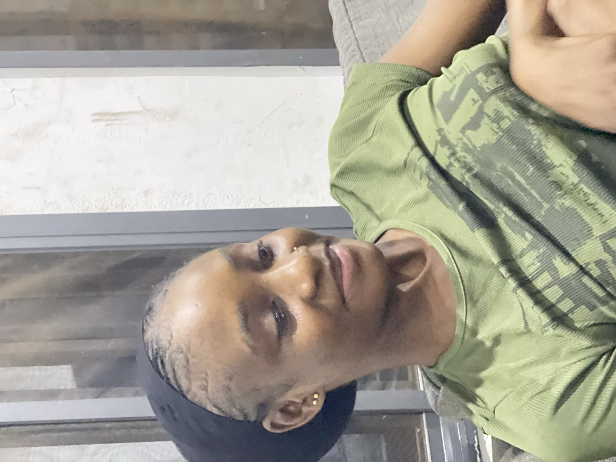 A young Black woman with braided hair, wearing a black headscarf and a green t-shirt with a graphic print, is seated on a grey sofa. She is looking downwards and to her right, with a slight smile. The background shows a window with vertical blinds and a textured, off-white wall. This setting is consistent with an indoor domestic environment in Abuja, Nigeria.