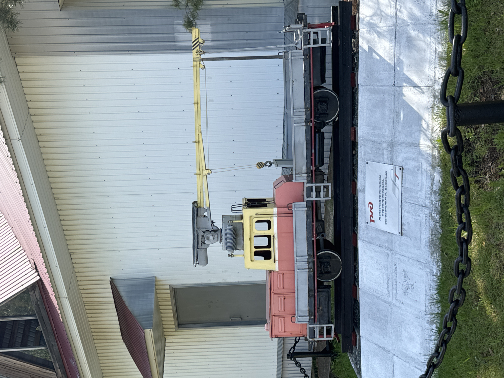 This image displays a static outdoor exhibit featuring a small, colorful rail-mounted vehicle, likely a railway crane or maintenance car. The vehicle has a yellow operator's cab, a reddish-orange main body section, and various grey and black mechanical components. A distinctive long, yellow crane arm with black and yellow striped warning signs extends upwards from the vehicle, equipped with hooks and cables. It rests on a short section of black railway track.
The exhibit is set on a paved area made of light-colored concrete slabs, bordered by green grass on the right. A thick black metal chain barrier runs along the right side of the paved area. In the background stands a building with light-colored, horizontal corrugated metal siding. Part of another building with a reddish-brown corrugated roof is visible on the far left.
Bright daylight illuminates the scene, casting distinct shadows from the vehicle and the chain onto the paved ground, suggesting a sunny day.
A white informational sign is placed on the concrete next to the rail vehicle. It prominently displays the red "РЖД" (RZD - Russian Railways) logo. Below the logo, Cyrillic text reads: "Железнодорожным основателям п. Высокий." This translates to "To the railway founders of the settlement Vysokiy," indicating the monument's purpose and its location context in Высокий, Russia. No people are present, and no activity is occurring beyond the static display.
