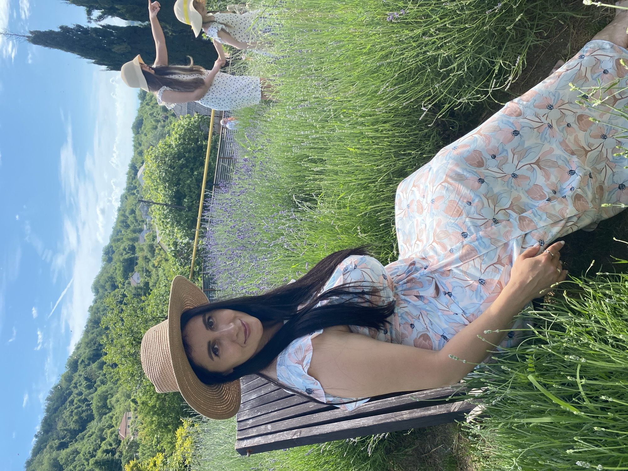 In Verkhneye Uchdere, Russia, a woman in a wide-brimmed straw hat and a floral dress sits on a dark wooden bench, facing the camera and smiling. She has long, dark hair and is surrounded by lush green grass and purple lavender. In the background, two figures, likely children, are visible in similar straw hats and white dresses. One points into the distance, while the other holds her hand. The scene is set against a backdrop of rolling green hills, with trees and buildings dotting the landscape under a bright blue sky with scattered clouds. The overall impression is one of a pleasant summer day in a rural or park-like setting.