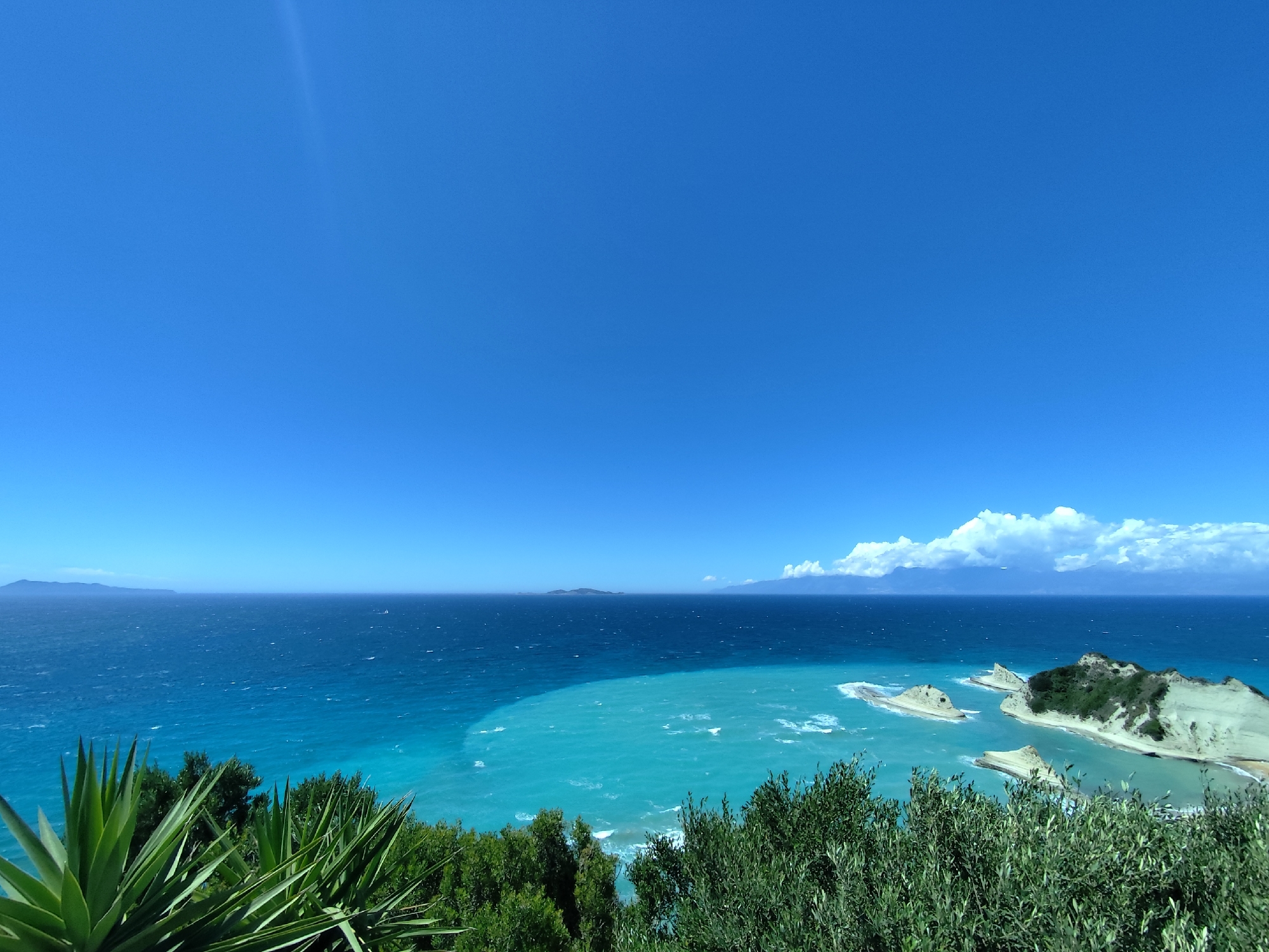 This panoramic photograph captures a stunning coastal landscape in Peroulades, Greece, under a brilliant, clear blue sky. The dominant feature is the expansive sea, which displays a remarkable two-tone effect: a deep, dark blue in the open water transitions sharply to a vivid, translucent turquoise closer to the shore.
Along the right side of the frame, rugged, light-colored cliffs and distinctive sea stacks emerge from the turquoise water, creating a dramatic coastline. These geological formations are topped with sparse green vegetation. In the far distance, faint landmasses, likely islands or distant coastlines, are visible on the horizon, with a scattering of white, cumulus clouds hovering above them on the right.
The foreground is framed by rich green vegetation, including spiky agave-like plants on the left and dense olive trees and shrubs on the right, suggesting the image was taken from an elevated vantage point overlooking the sea. The scene is bathed in bright, clear daylight, indicating it is likely midday or early afternoon. The weather appears sunny and calm, emphasizing the vibrant natural colors. There are no people visible, nor any discernible text in the image.