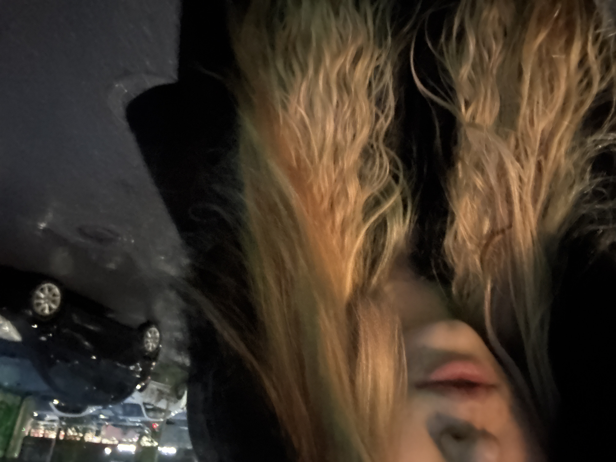 The media file is a close-up, vertically oriented photograph of a person's face and hair, with a car and other background elements reflected in a wet surface. The primary subject is the person, whose face is partially visible, with their lips, nose, and cheek in focus. They have long, wavy hair with streaks of blonde, brown, and possibly some green or yellow tones. The person appears to be looking down or away from the camera.
Reflected in the wet surface, which seems to be a street or a similar dark, glossy ground, is a black car. The car's wheels and the general shape are discernible. Further in the reflection, other cars and some blurry lights suggest an urban or roadside setting, possibly a parking lot or street at night.
The overall lighting suggests it is dark, and the wet surface implies it has recently rained or is raining, creating reflections. The image has a somewhat dramatic and moody feel due to the lighting and reflections. There is no visible text in the image. The activity is not clearly defined, but the pose of the person and the environment suggest a moment captured, perhaps during a walk or while waiting.