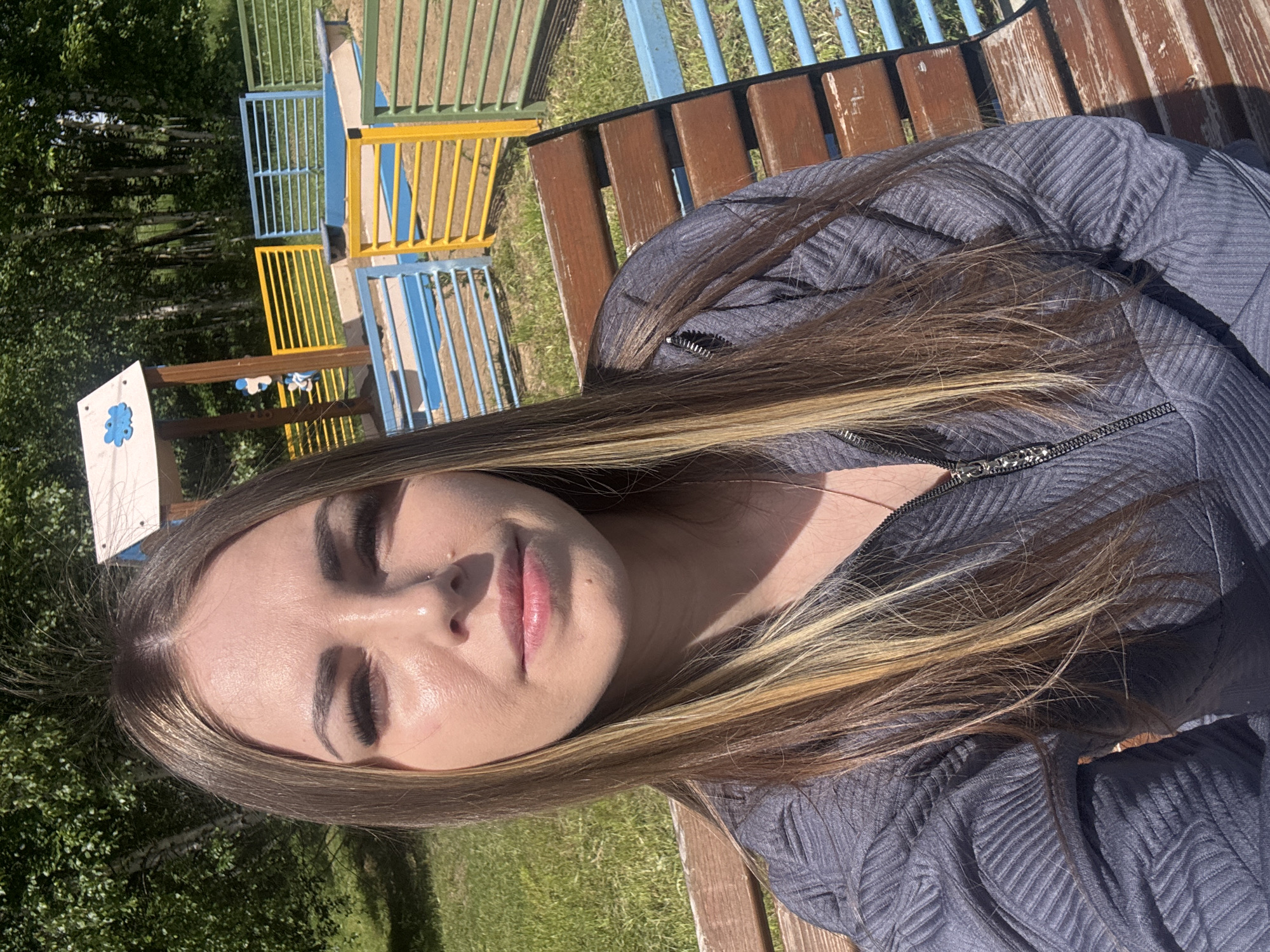 A woman with long brown and blonde hair is seated on a wooden bench in an outdoor area. She is wearing a gray jacket with a zipper. In the background, there is a playground structure made of blue and yellow metal bars, and a sign with a blue emblem. The setting appears to be a park or recreational area with grass and trees visible. The exact location within Shevlyakovo, Russia cannot be determined.