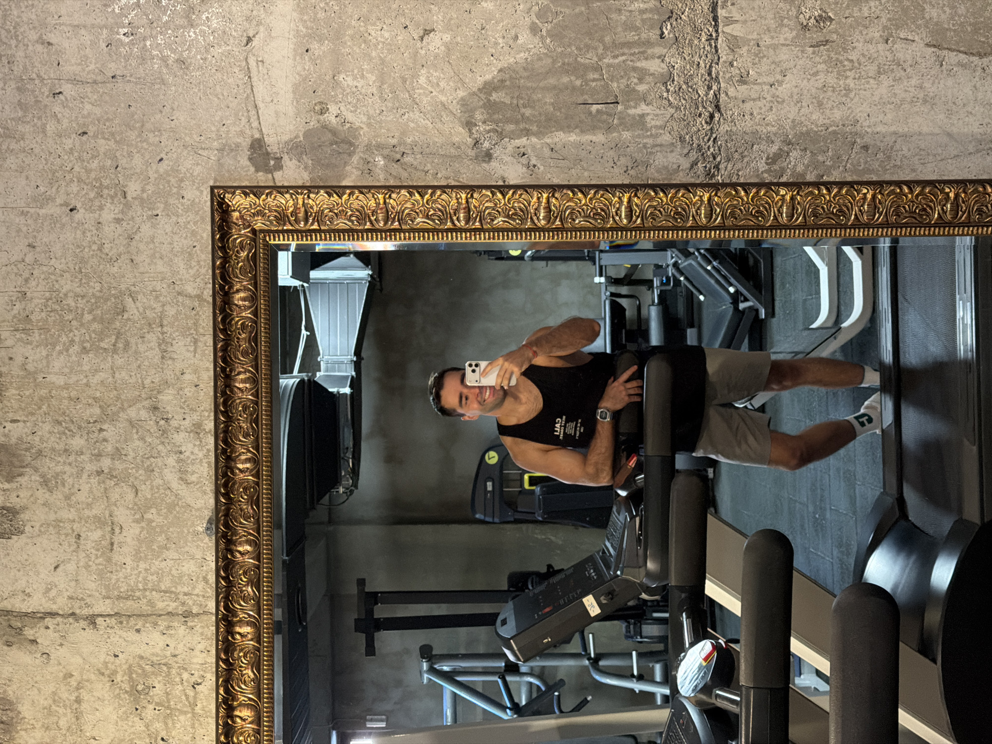 A man is reflected in an ornate gold-framed mirror affixed to a concrete wall. He is fair-skinned with dark hair, wearing a black tank top, gray shorts, and white socks. He is holding a white smartphone and taking a selfie in what appears to be a gym. Weightlifting equipment, including machines with adjustable seats and levers, is visible in the background. The gym is located in Tbilisi, Georgia.