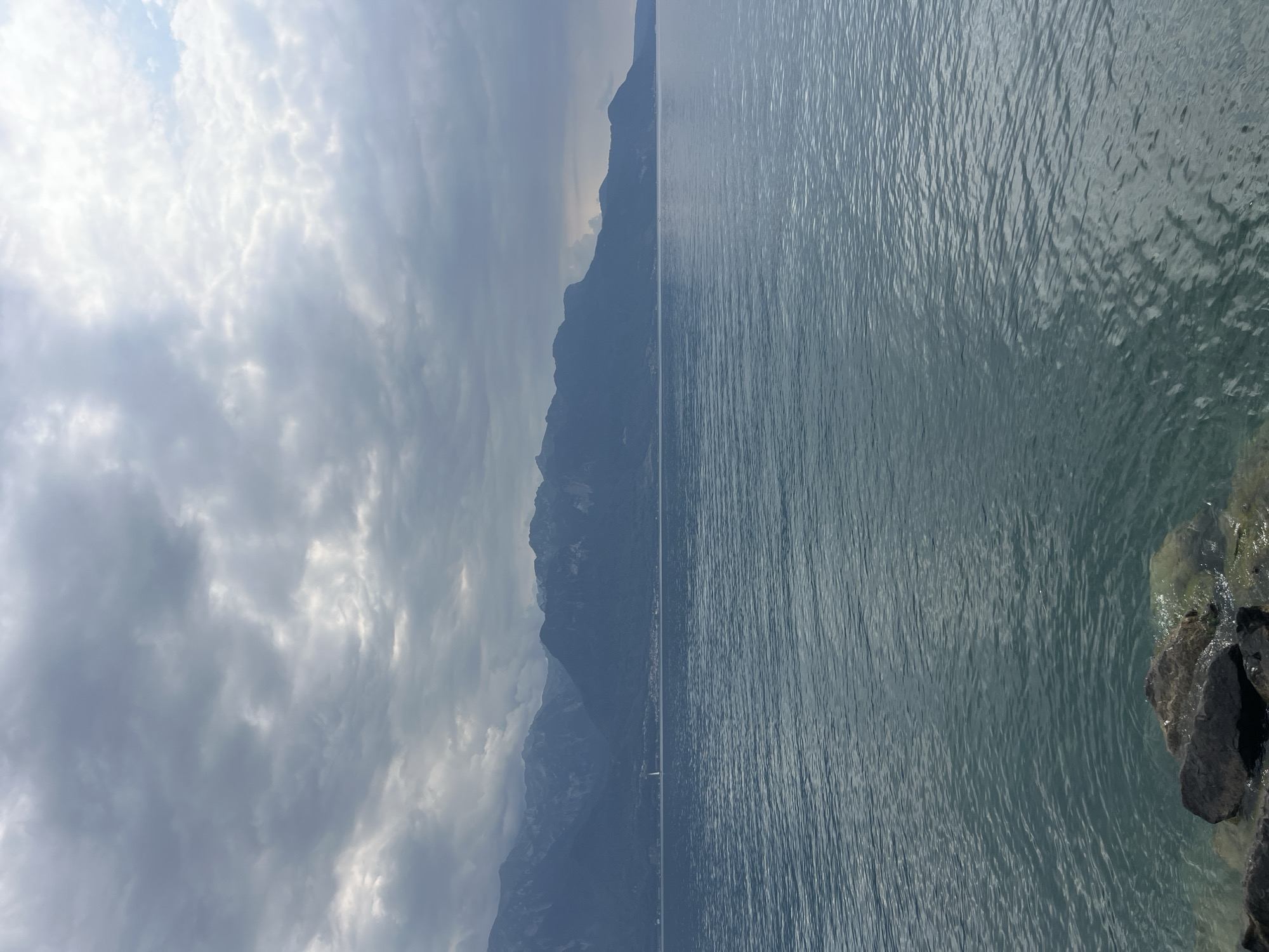 An exterior, medium shot captures a wide expanse of water under a cloudy sky, with a mountainous landscape in the background. The water, a deep teal color, is rippled, suggesting a gentle breeze. In the foreground, dark, wet rocks are visible at the edge of the water, indicating a shoreline. The mountainous terrain behind the water is shrouded in a hazy, bluish-gray hue, with some hints of greenery on their slopes. The sky is filled with heavy, gray clouds, with patches of lighter sky peeking through in some areas, giving the scene a somewhat somber or dramatic atmosphere. A small, white sailboat is visible in the distance, adding a point of interest to the vastness of the lake and mountains. This vista is characteristic of the scenery surrounding La Tour-de-Peilz, Switzerland, particularly along the shores of Lake Geneva, with the Alps forming the dramatic backdrop.