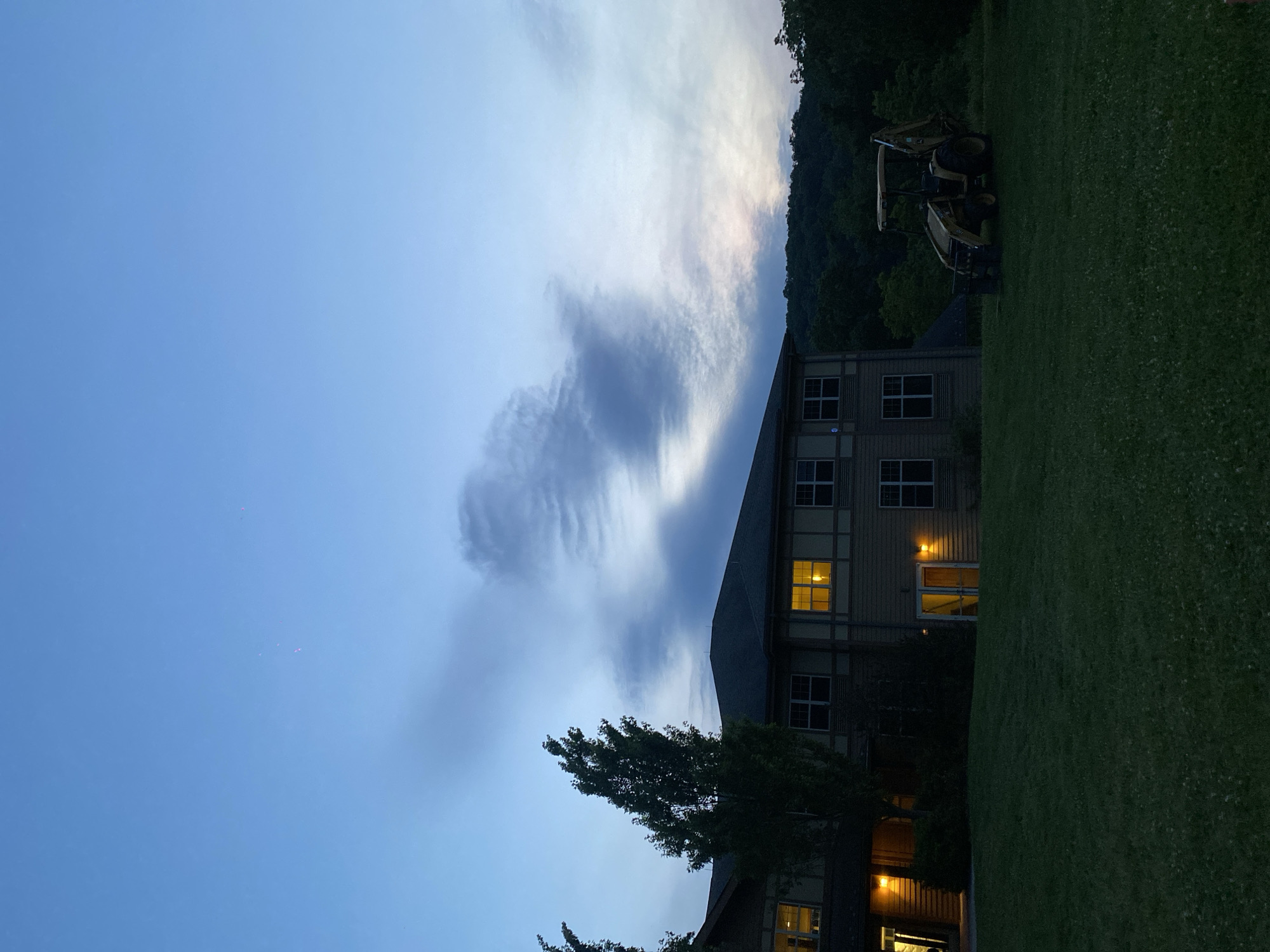 This image depicts a dusk scene at an apartment complex located in Washington Township, United States of America. The sky is a gradient of blue and purple, with wispy clouds. In the foreground, a well-maintained lawn is visible, leading up to a two-story building with several lit windows, indicating interior activity. The building features a light-colored facade with darker trim and numerous windows. To the right of the building, a yellow backhoe loader is parked on the grass. A dense line of trees borders the property in the background, suggesting a semi-rural or suburban setting. The overall atmosphere is peaceful and quiet, with the fading light casting long shadows across the scene.