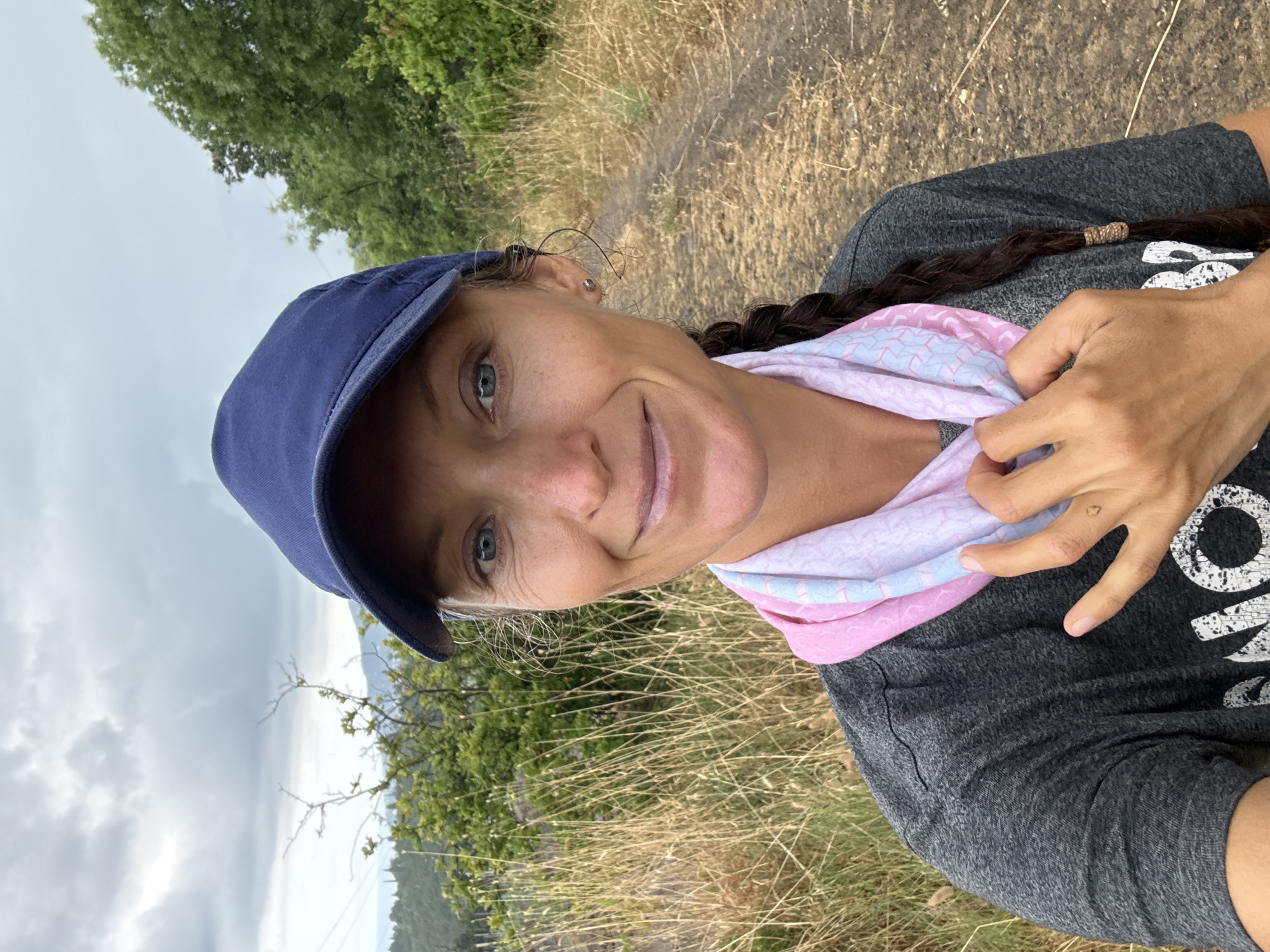 A woman wearing a blue baseball cap and a grey t-shirt is standing outdoors. She has a pink and blue bandana around her neck. The woman is smiling and looking at the camera. Her hair is in a braid. The background appears to be a trail in a natural setting with dry grass and trees.