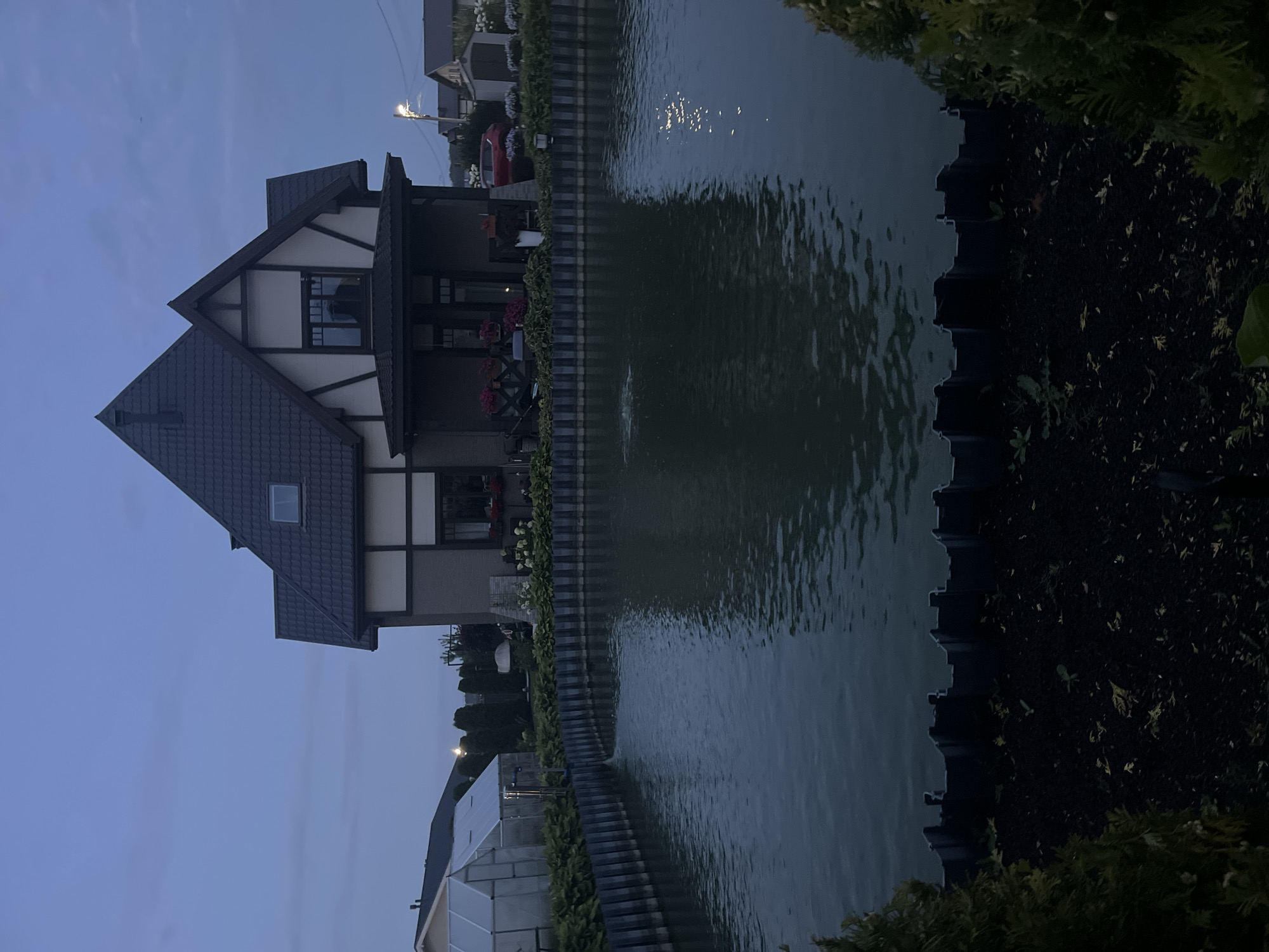 This is a photograph taken at dusk in the Babigonskaya Valley, Russia, featuring a two-story house with a dark tiled roof and decorative half-timbering. In front of the house is a pond with a black retaining wall around its perimeter. The pond has ripples on its surface, and a small fountain is visible in the center. To the left of the house, there is a greenhouse. Various trees and shrubs are present in the landscape, including some evergreen trees in the foreground on the right and left edges of the frame. A red car is parked in front of the house. The sky is a deep blue, indicating late evening or early morning. The overall scene depicts a residential property with a pond and surrounding garden.