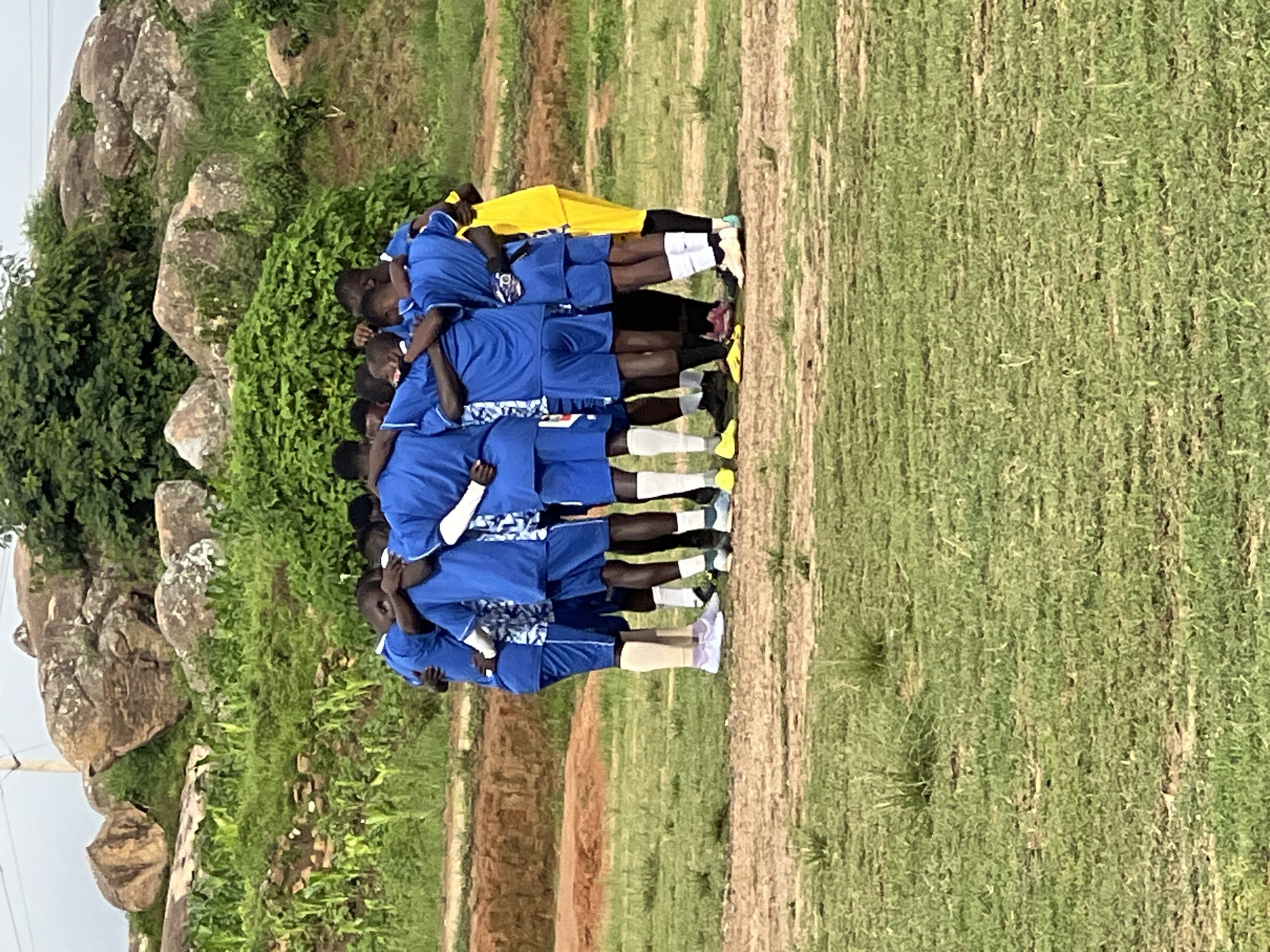A group of young men, likely a soccer team, are huddled together in a circle with their arms around each other's shoulders. They are wearing matching blue jerseys with white and blue patterned accents and blue shorts. Some are wearing white socks with colored athletic shoes, while others are wearing dark socks and shoes. One person on the edge of the huddle is wearing a bright yellow shirt and pants. They appear to be on a grassy field with patches of dirt, possibly a soccer pitch. In the background, there are large rocky outcrops and green vegetation, suggesting a natural, somewhat rugged environment. The sky is overcast. This scene is consistent with the landscape found in and around the city of Jos, Nigeria, known for its rocky terrain and hilly topography.