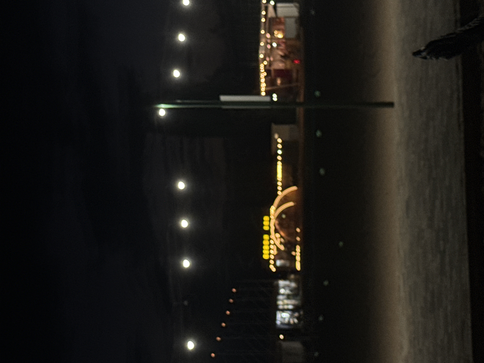 The image captures an outdoor scene at night, likely in a recreational or event space, possibly near Issyk-Kul Lake in Sary-Oy, Kyrgyzstan, given the context of the location. In the foreground, the ground appears to be a dirt or gravel surface. A dark, indistinct object is visible in the lower right corner.

The midground features a pole with lights on top, positioned slightly off-center to the right. Behind this pole, several structures with illuminated string lights are visible. One structure appears to be a building with lights in windows and along its roofline. Another prominent feature is a large, arched structure, possibly a tent or tunnel, also adorned with numerous yellow lights, suggesting it's a decorative element or entrance. Several smaller, unidentifiable structures with lights are scattered in the background.

The sky is dark, with a few scattered lights, likely from overhead fixtures or distant sources. The overall impression is of a nighttime gathering or festival, with artificial lighting creating a festive atmosphere against the dark backdrop. The image is slightly out of focus, which gives the lights a soft, blurred quality.