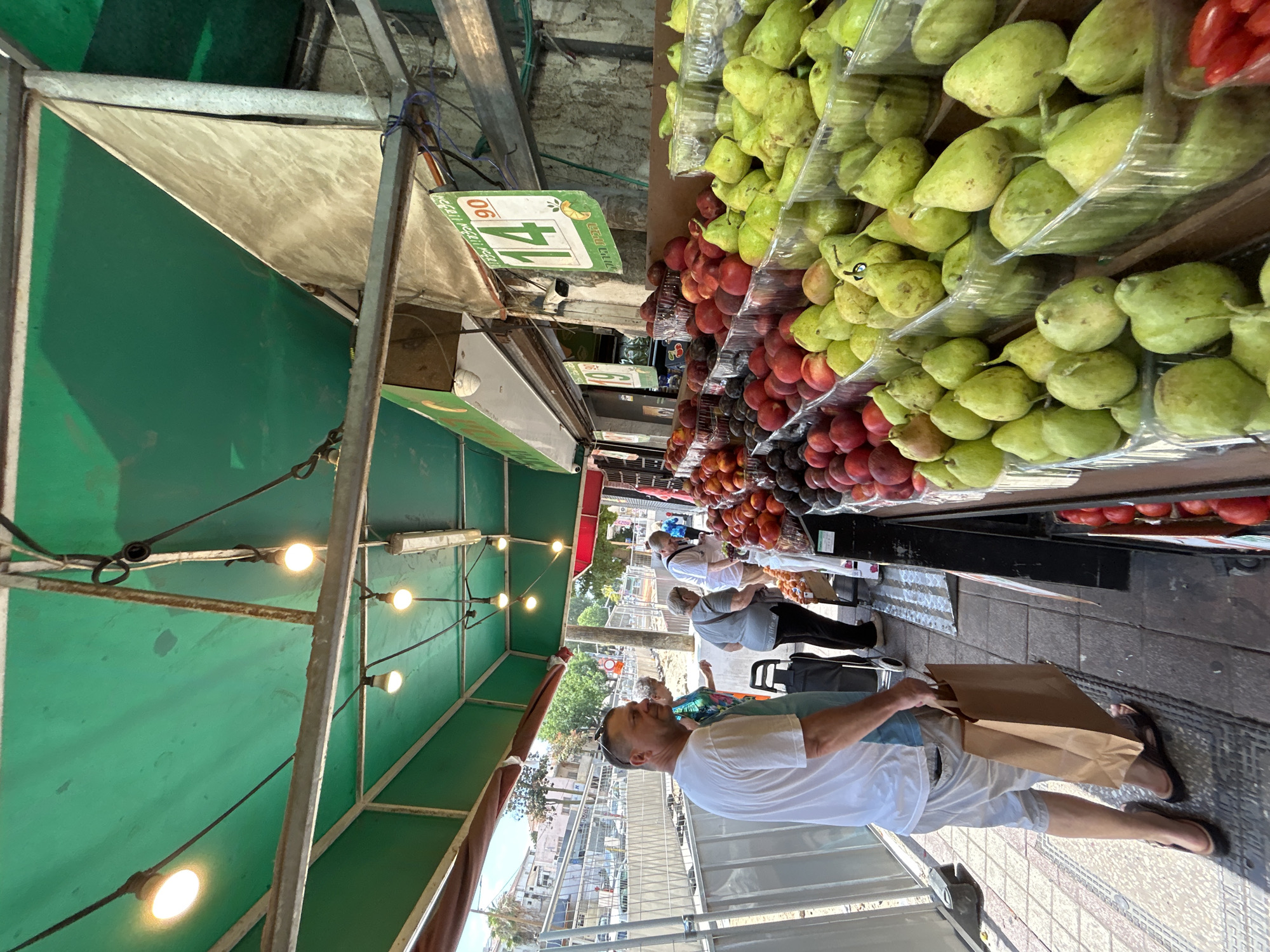 A street market scene in Rishon LeZion, Israel features a man in a white t-shirt and white shorts carrying a brown paper bag. He is walking on a sidewalk past a fruit stand. The fruit stand displays a variety of fruits, including pears, peaches, and plums, in clear plastic containers. Some of the fruits have price tags, one indicating "14.90". Behind the fruit stand, a row of buildings is visible, and further in the background, construction fencing and equipment suggest ongoing urban development. The market is covered by a green awning, with lights hanging from the structure. Several other people are browsing the fruit stand. The scene captures a typical urban market environment with a focus on fresh produce.