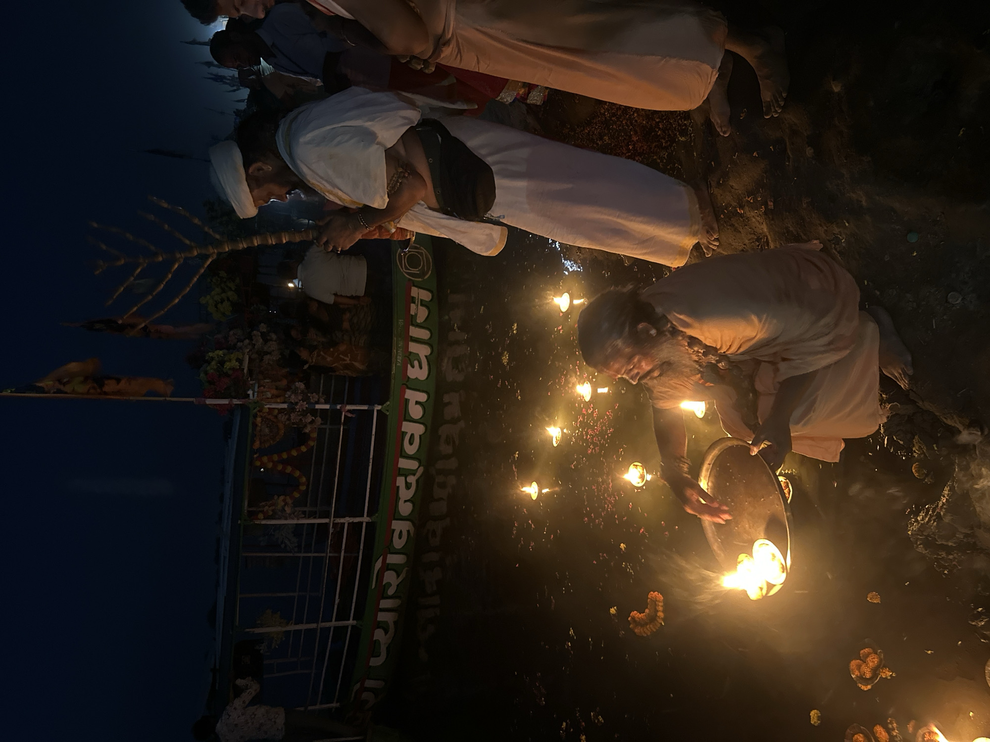 At night in Vrindavan, India, people are gathered on what appears to be a riverbank for a religious ceremony. One individual kneels and holds a tray with lit flames over the water. Flowers float on the water, illuminated by the candles and flames. Others, in white robes, are standing or kneeling in the foreground, also involved in the ritual. In the background, a structure is adorned with garlands and religious symbols. The text "प्यारो वृंदावन धाम" is visible on the side of the structure. The scene is dimly lit by the candlelight.