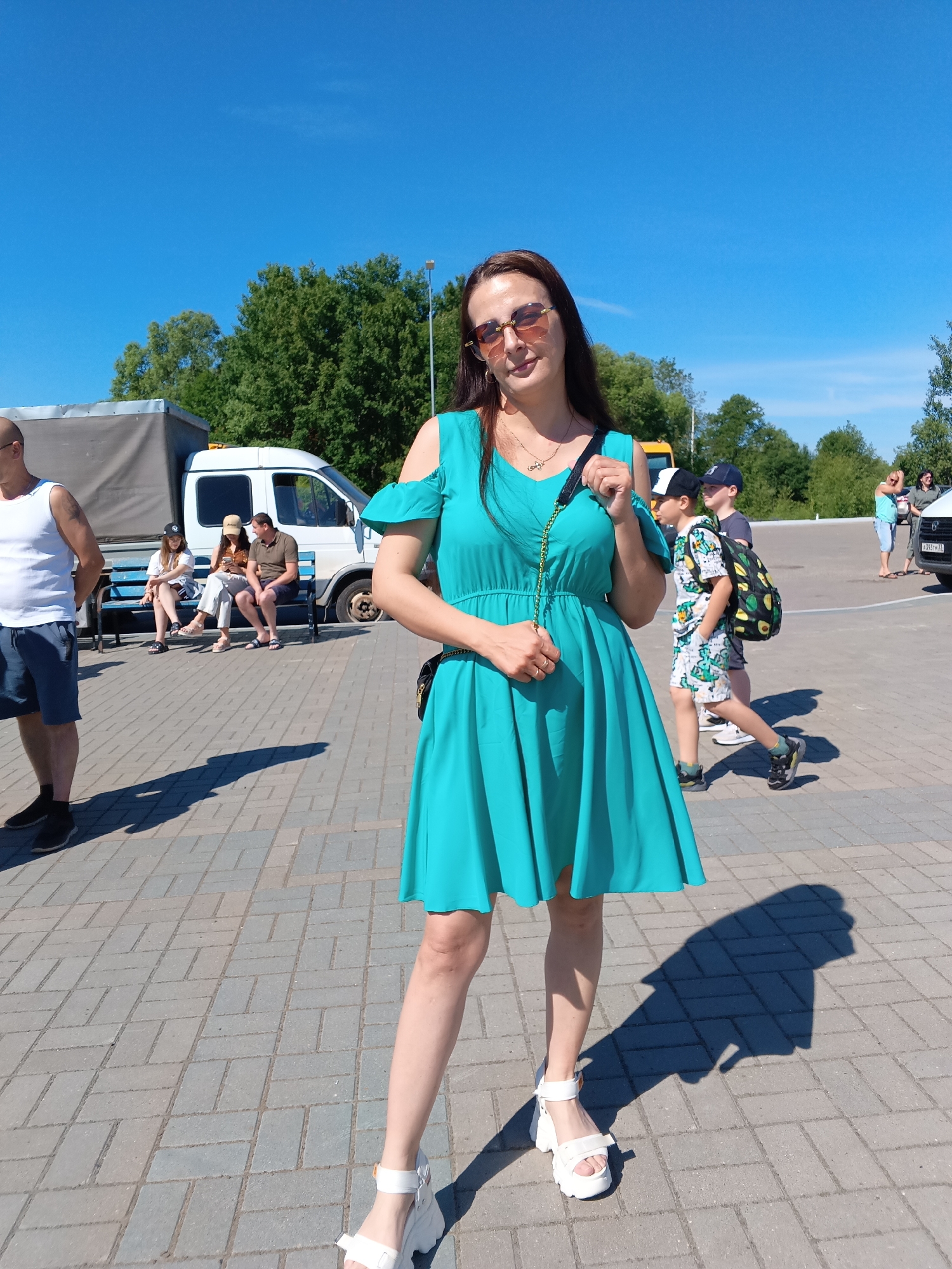 This image captures an outdoor scene in Sudogda, Russia, on a bright, sunny day, indicated by the clear blue sky and sharp shadows. The ground is paved with grey rectangular pavers.
In the immediate foreground, a woman with long, dark brown hair and sunglasses is centered, facing the viewer with a slight smile. She is wearing a teal, V-neck, cold-shoulder dress that extends above her knees, and white platform sandals. A thin gold chain necklace with a small pendant is visible. She holds the strap of a small black bag with a gold chain accent in her right hand. Her shadow is distinctly cast to her right.
In the midground, from left to right:
A bald man in a white tank top, dark shorts, and dark shoes stands with his left side partially facing the viewer. Behind him, a grey truck with a tarp cover is parked. Adjacent to the truck, a white passenger van is parked next to a blue metal bench. On the bench, two individuals are seated; one person wears a light-colored hat and appears to be looking at a device, while another man in a dark polo shirt and shorts sits beside them. Further right, two boys walk; the closer boy wears a white and black baseball cap, a patterned light-colored t-shirt and shorts, and a black backpack adorned with green avocado patterns. He wears sneakers. Another boy in a dark cap and t-shirt is partially visible behind him.
In the distant background, several green trees form a dense line against the clear blue sky. Additional indistinct figures and parked vehicles are visible on the far right, including a car with a Russian license plate (B393TH 33). A street lamp pole is also visible on the far right.