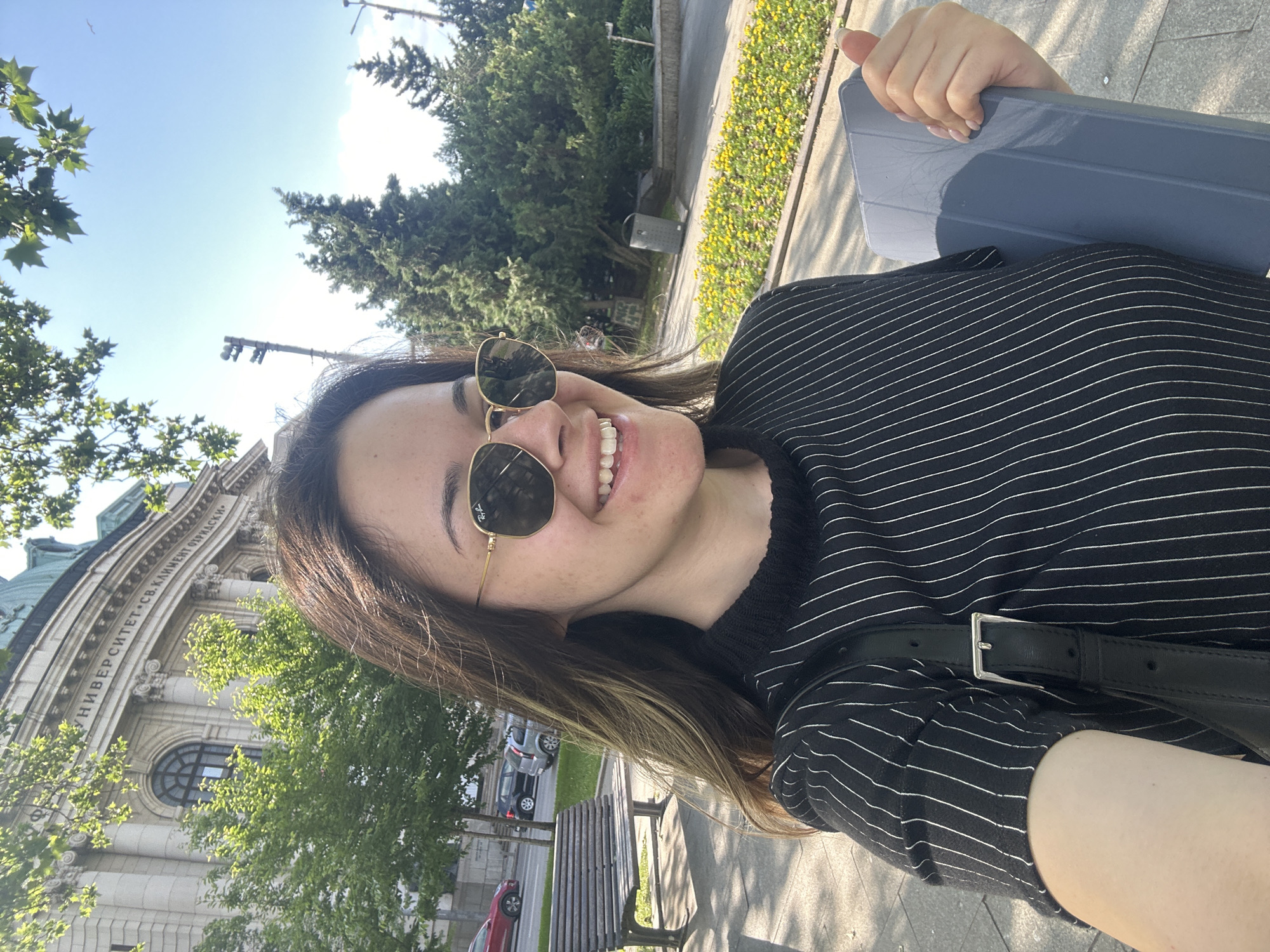 A young woman with shoulder-length brown hair and wearing round sunglasses is posing for a selfie outdoors. She is smiling, wearing a black and white striped shirt, and holding a gray tablet in her hand. In the background, there is a large building with a dome and classical architecture, identified as the Sofia University "St. Kliment Ohridski." There are also trees and a bench visible, suggesting a park or public space adjacent to the university. The location is in Sofia, Bulgaria.