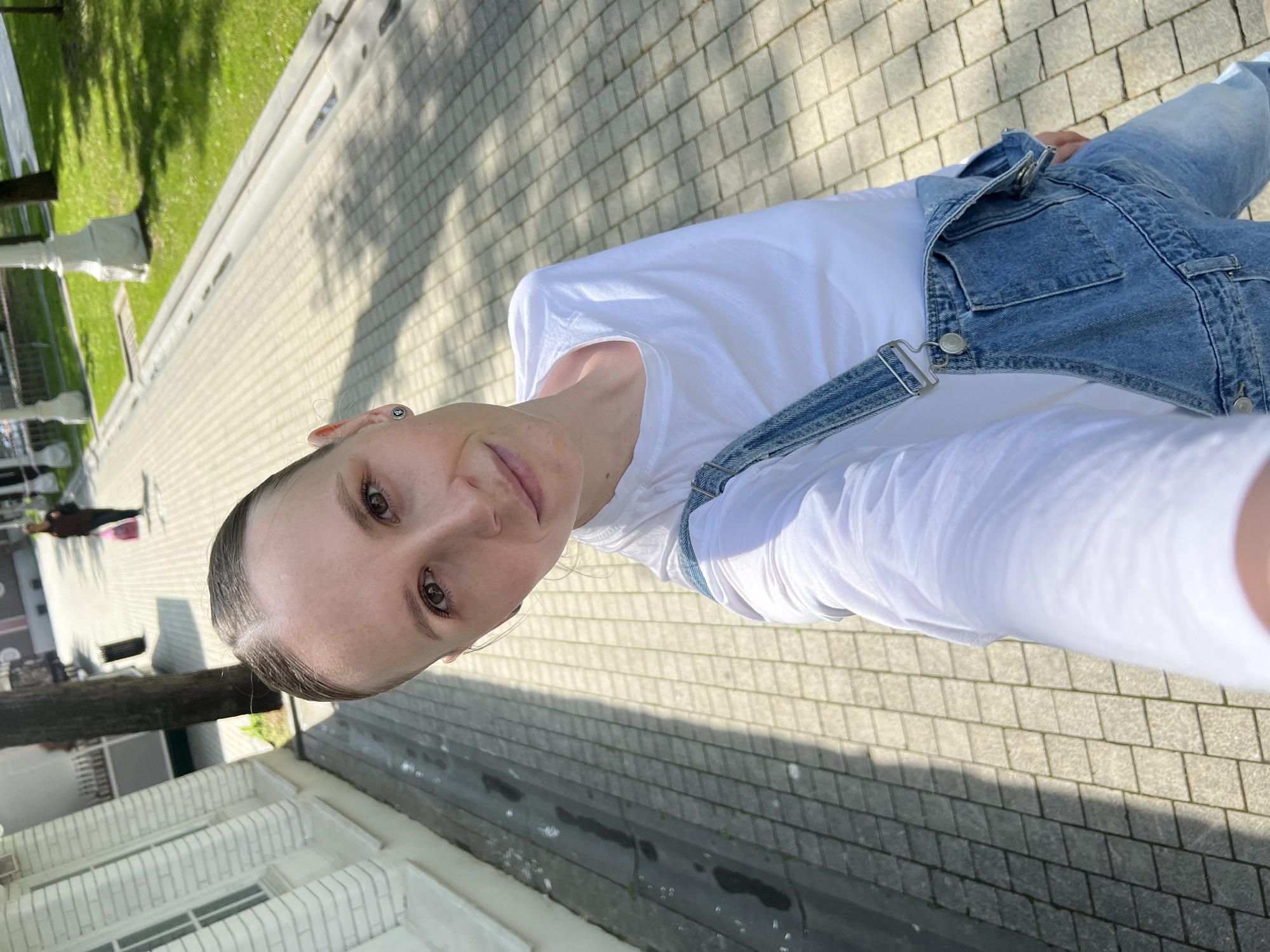 A selfie captures a person wearing light blue denim overalls over a white long-sleeved t-shirt, standing on a gray brick walkway. Visible behind and to the left of the person is a white building with visible windows and a grassy area with trees. Further away, blurred figures and buildings are visible. The photograph was taken on a sunny day, evidenced by the shadows cast on the brick walkway. It is not possible to confirm the exact location within Moscow, Russia from the image alone.