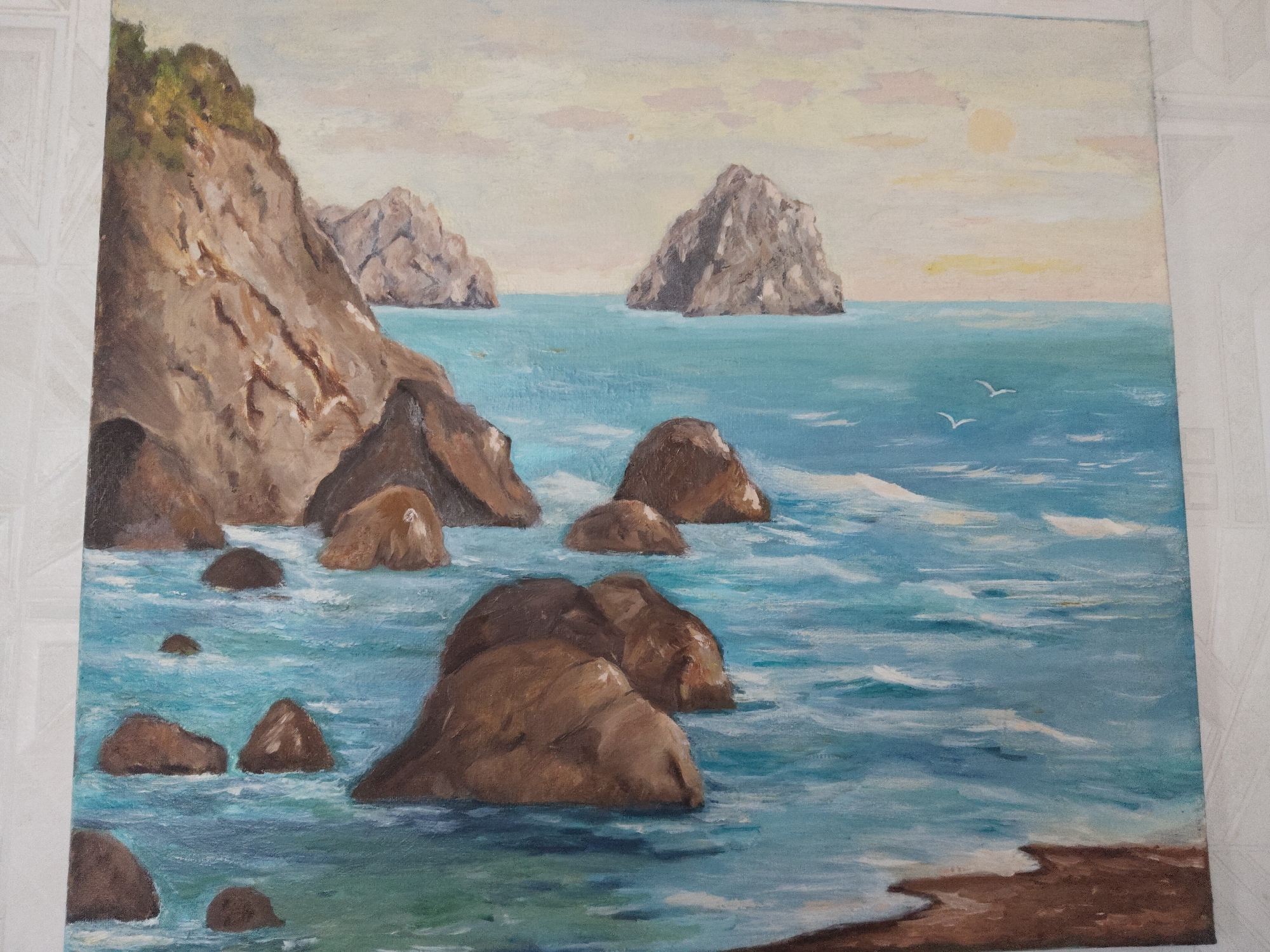 The image is a painting depicting a coastal scene with rocky formations. The foreground features numerous brown rocks of varying sizes emerging from the turquoise and blue water, which is rendered with visible brushstrokes to suggest movement. A prominent, large rock formation dominates the left side of the painting, topped with green vegetation. In the distance, three more rocky outcrops rise from the sea. The sky is a muted yellow and pale blue, indicating either sunrise or sunset. Two white birds are depicted flying in the distance, adding a touch of life to the scene. The overall style is reminiscent of impressionism. The location and city are not provided, unknown.