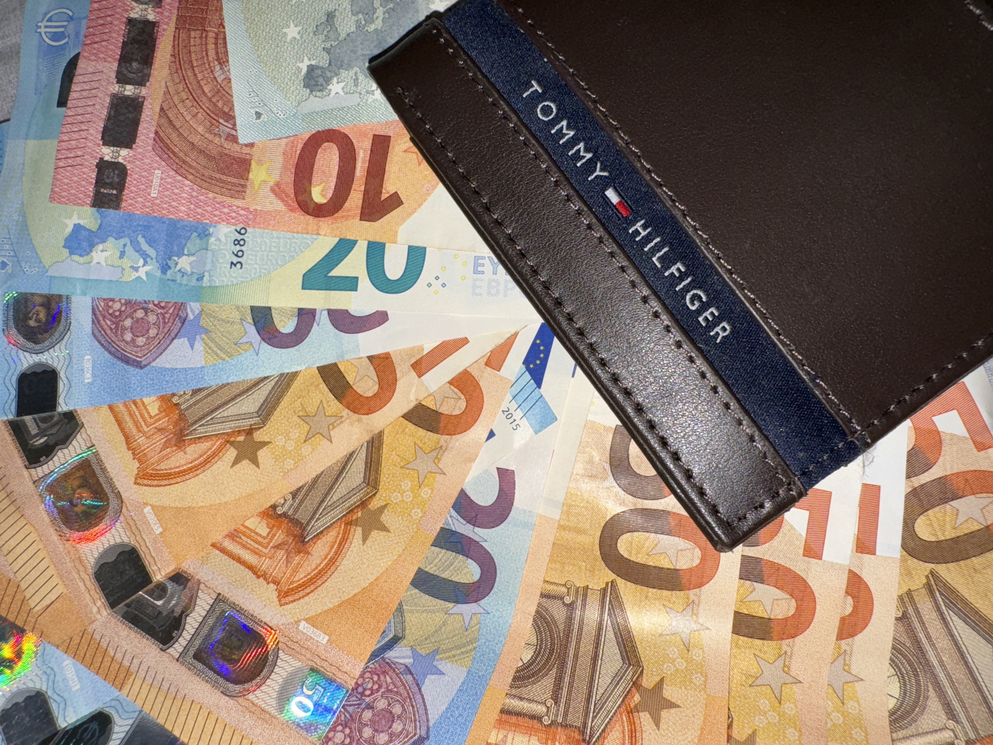 The image shows a brown Tommy Hilfiger wallet placed on top of a fan of Euro banknotes. The banknotes are primarily 50 Euro notes, with some 10 and 20 Euro notes also visible. The location of the image is unknown.