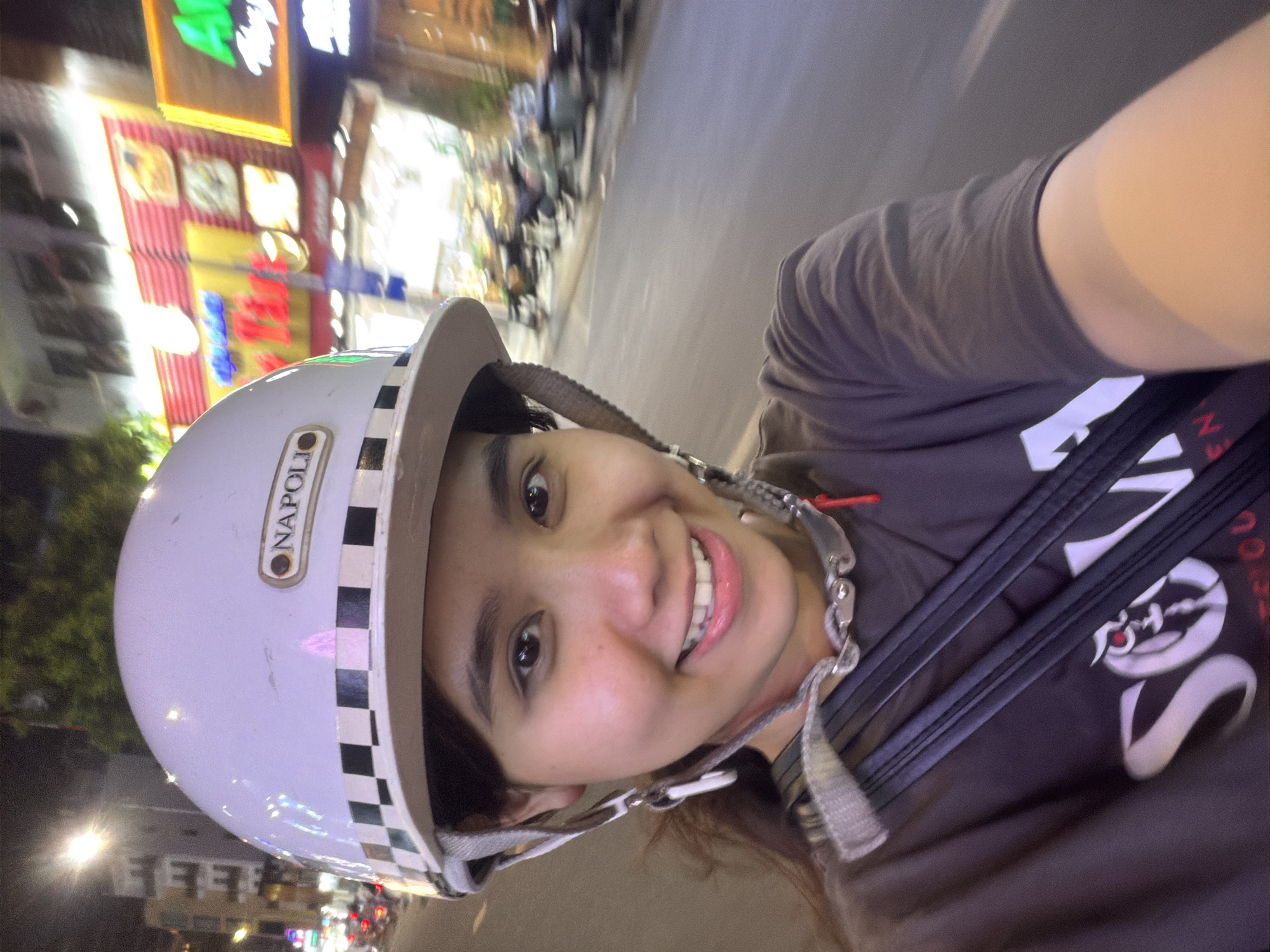 A young woman takes a cheerful selfie while likely riding or being a passenger on a scooter or motorcycle at night in Đà Nẵng, Vietnam. She is positioned close to the camera, looking directly at the viewer with a broad, genuine smile that highlights her teeth. Her dark eyes are bright, and her cheeks show a light blush.
She wears a white half-face helmet with a white and black checkerboard pattern along the rim and the brand name "NAPOLI" in a small oval logo on the side. The helmet's chin strap is visible. Her attire includes a dark grey or brown t-shirt with some stylized white lettering, too blurry to fully decipher, on the front. A dark strap, possibly from a bag or another piece of gear, crosses her chest.
The background is an urban street scene at night, characterized by significant motion blur, especially in the upper and far background, suggesting movement. Brightly lit commercial signs, featuring various colors like red, yellow, green, and blue, illuminate the scene, casting a glow. Other blurred lights and shapes indicate buildings and possibly other vehicles or pedestrians. The overall ambiance is lively and nocturnal.