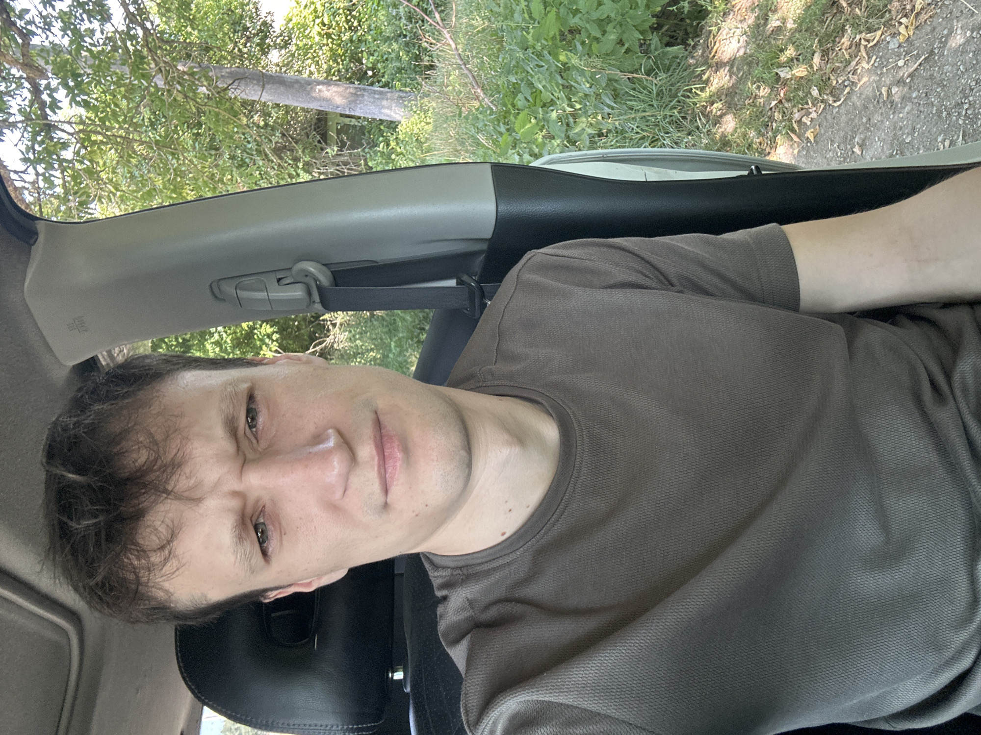 A man with brown hair and a questioning expression sits in the driver's seat of a car, looking at the camera. He is wearing a brown t-shirt. The interior of the car is visible, including a black seat and a grey pillar with a seatbelt. Outside the car, lush green trees and foliage are seen. The scene appears to be in a rural area, possibly on a dirt road. The location is specified as Makiv, Ukraine.