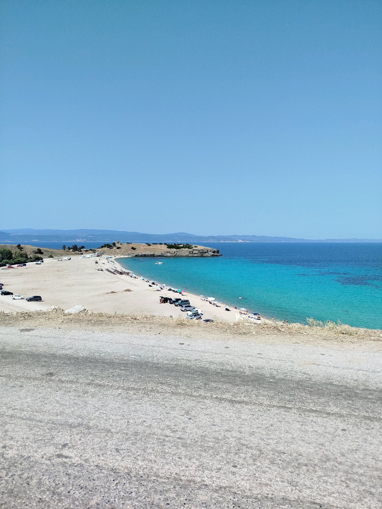 The image depicts a scenic view of a beach located in Sithonia Municipal Unit, Greece. The scene is dominated by a vast expanse of clear blue sky, with a gradient of lighter blue towards the horizon. Below the sky, a tranquil turquoise sea meets a sandy beach, which curves along the coastline. The beach is dotted with various beach umbrellas, some empty and others with people relaxing underneath. Several beachgoers can be observed on the sand and in the shallow waters.
Adjacent to the beach, a dirt road serves as a parking area for a number of cars. Some of these vehicles are parked near the water's edge, while others are positioned further back along the road. In the distance, a hilly coastline with sparse vegetation stretches across the horizon, creating a picturesque backdrop. A small white boat is visible on the water, adding a touch of activity to the otherwise serene seascape. The foreground of the image is occupied by a rough, grey asphalt surface, suggesting a vantage point from a road or elevated area overlooking the beach. The overall impression is one of a popular summer destination, bathed in bright sunlight.
