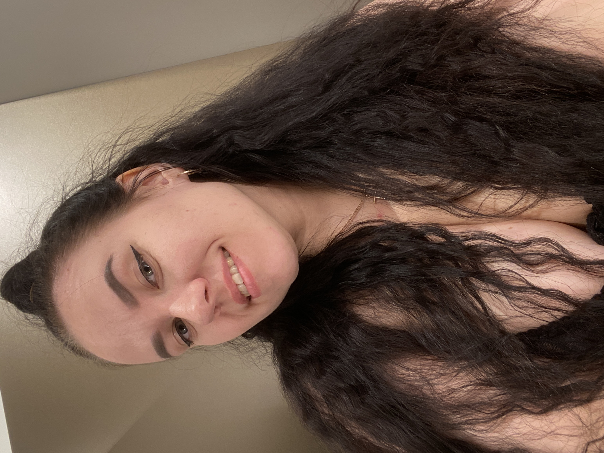 A woman is posing for a selfie. She has long, dark hair and is smiling. She has eyeliner on her eyes and an earring in her left ear. She is wearing a gold necklace. Her skin tone is fair. A beige wall is in the background. The city of Perm, Russia is not visually discernible in the image.