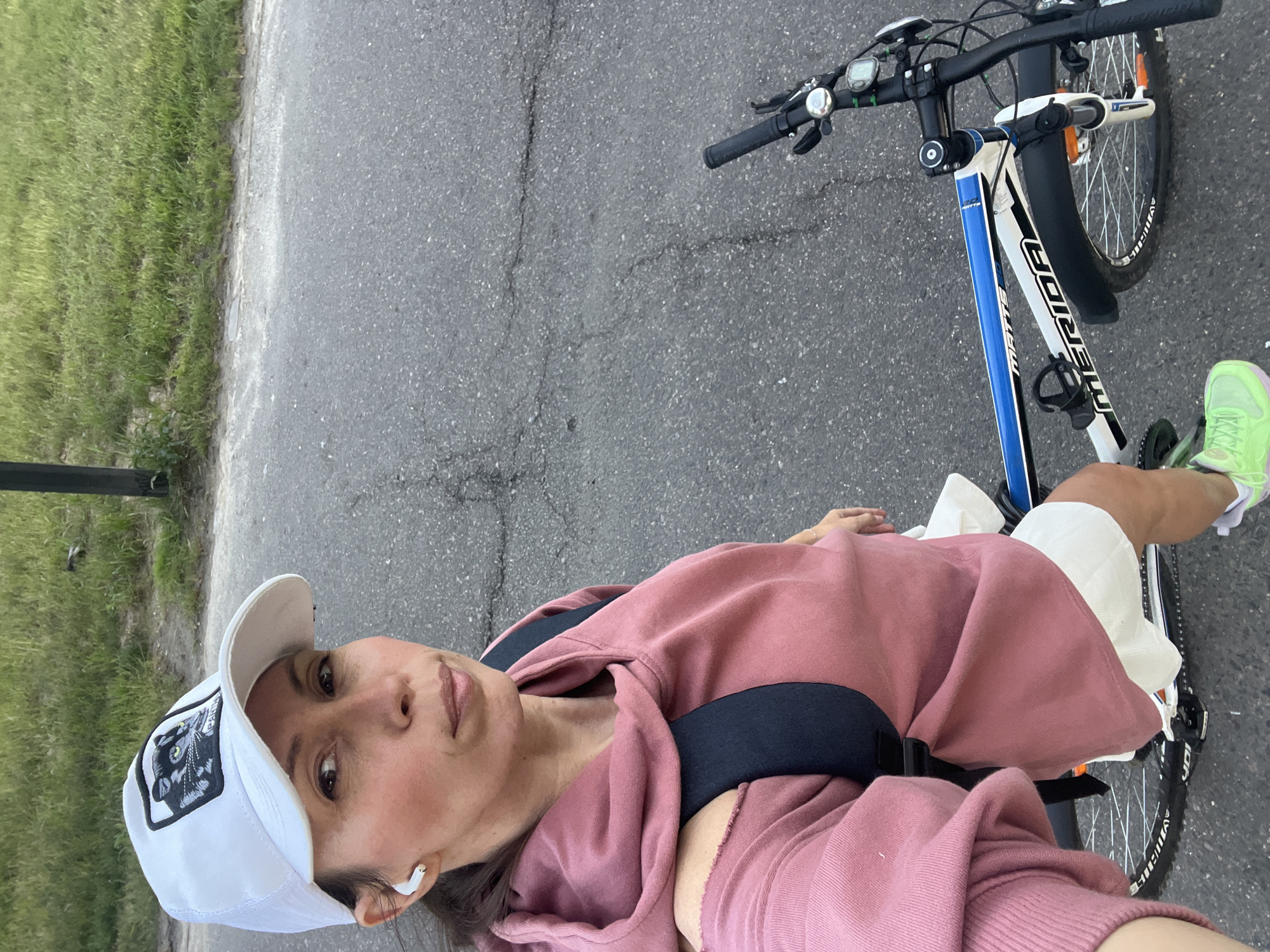A female subject, appearing to be in her 30s to 40s, is captured in a selfie-style photograph outdoors in Moscow, Russia. She wears a white baseball cap featuring a black patch with an illustration of a black panther's head with prominent green eyes. A white wireless earbud is visible in her right ear. Her attire includes a dusty rose-colored hooded sweatshirt with cut-out shoulder details and white shorts. A dark-colored backpack is worn on her back. She wears bright neon green athletic shoes with purple soles. The subject is positioned astride a Merida Matts bicycle, which has a white and blue frame with black lettering and components, including a black front fender. The setting is an asphalt road or pathway exhibiting cracks and small aggregate. Adjacent to the asphalt, a strip of green grass and weeds runs alongside a dark, vertical pole. The scene is illuminated by natural daylight.