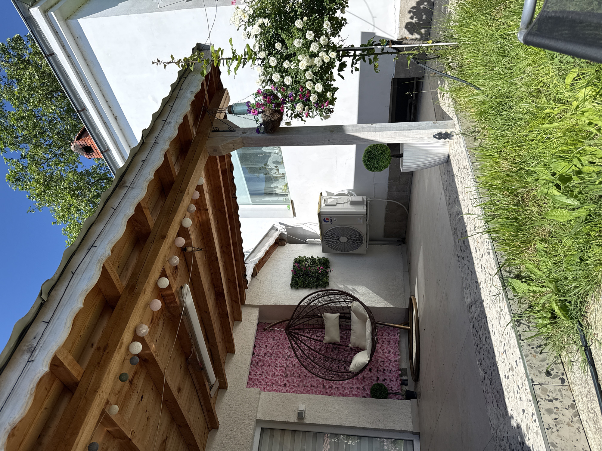 The image depicts an outdoor patio space. A wooden pergola with string lights provides partial shade. A hanging chair with white cushions sits against a backdrop of pink artificial flowers. A window is visible beneath the backdrop. An air conditioning unit is mounted on the wall above the chair. A square arrangement of artificial greenery is located above the chair. Potted flowers hang above the patio. Green grass lines the patio edge. The patio is located in Elhovo, Bulgaria.