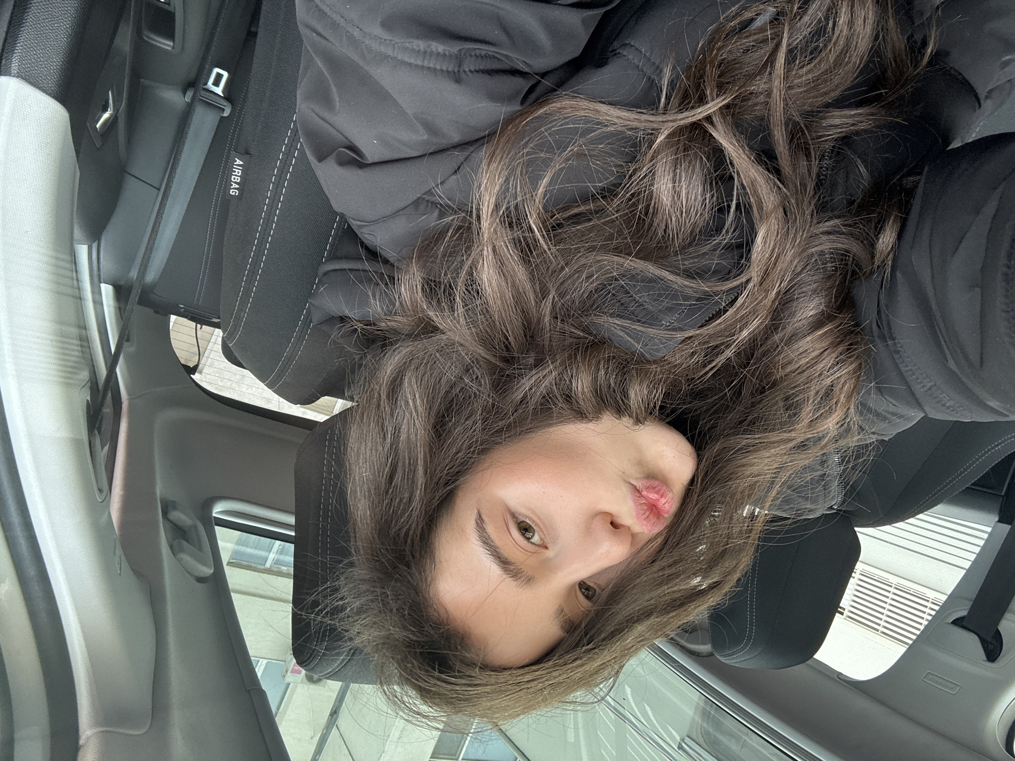 A woman is inside a car, possibly in the driver's seat or passenger seat. She has long, wavy brown hair and is wearing a black puffy jacket. She is making a kissing face toward the camera. The car interior is visible, including the seat, seatbelt, and airbag label. Outside the car window, a building or structure can be seen. The location appears to be a city but cannot be definitively determined.