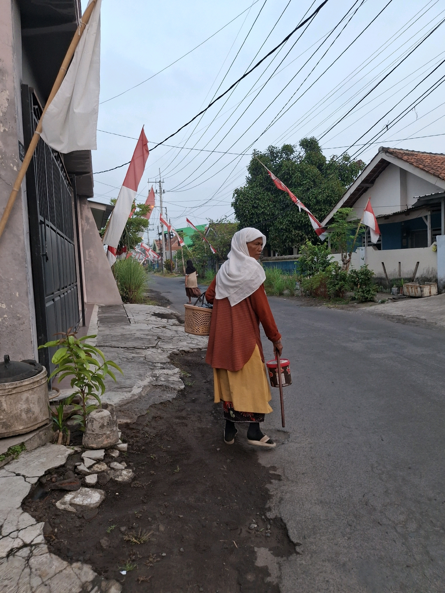 The image depicts a street scene in Lumajang, Indonesia, adorned with Indonesian flags. In the foreground, a woman wearing a white hijab, an orange cardigan, and a yellow skirt walks on the side of the road, holding a walking stick and a cylindrical container. The street is paved with asphalt and has some dirt and debris on the side. Buildings with tiled roofs line the street, and lush green trees are visible in the background. Electrical wires are strung across the sky, indicating an urban or semi-urban setting. The overall atmosphere suggests a normal day in a residential area, possibly during a national celebration given the presence of flags.