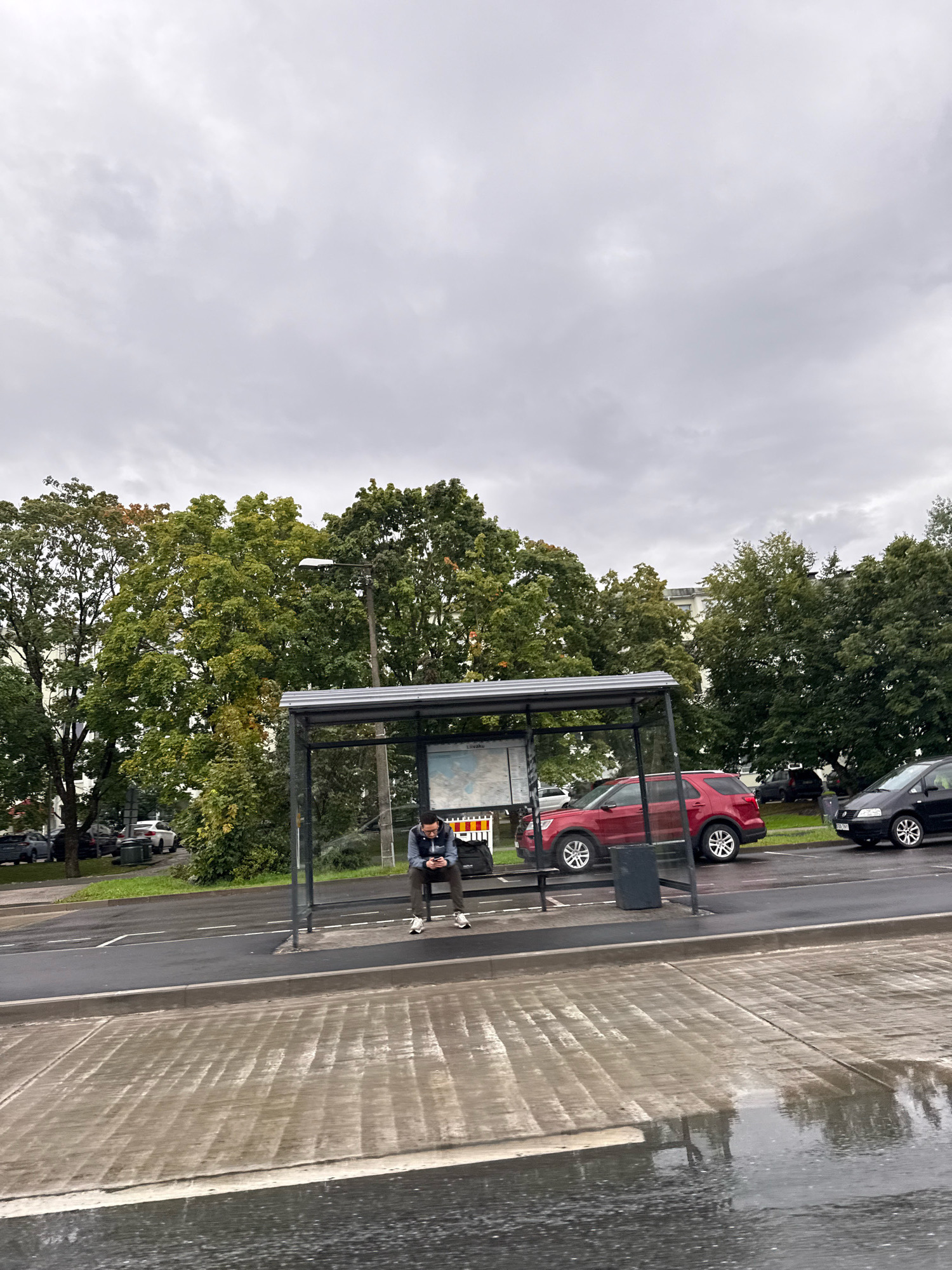 A man is sitting alone at a bus stop on a cloudy day in Tallinn, Estonia. He is wearing a grey hooded sweatshirt and dark pants, and is looking down at his phone. The bus stop has a metal roof and glass panels, and there is a map displayed inside. There are cars parked along the street behind the bus stop, and trees line the background. The ground in front of the bus stop is wet and reflects the surroundings, suggesting it has recently rained.