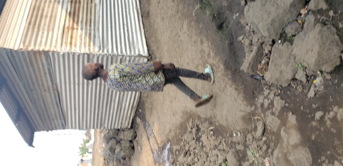 The image depicts a daytime scene in Buzove, Democratic Republic of the Congo. A young individual with short hair, wearing a multi-colored patterned long-sleeved shirt and dark trousers, is walking right-to-left across the frame on a dirt path. The person is wearing light blue open-toed sandals. The ground is uneven, composed of packed dirt, loose stones, and areas with standing water or damp soil. To the left of the path, a collection of dark, irregular rocks or rubble is present. In the background, a structure constructed from corrugated metal sheets with a sloped roof is visible, partially obscured by the individual. Further in the background, out of focus, appear to be more structures and some sparse vegetation under an overcast sky.
