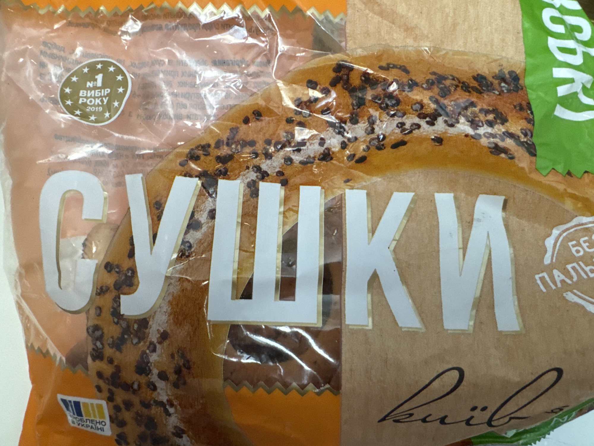 The image displays a close-up of food packaging. The packaging is primarily orange and brown with some green and white accents. Large, white, capitalized letters with gold trim spell out "СУШКИ" which translates to "Sushki" (dried bread rings) in English. To the left of the text, there is a circular seal with stars and the text "№1 ВИБІР РОКУ 2019" which means "No. 1 CHOICE OF THE YEAR 2019." To the right of the main text, there is a green label with white text that appears to be "БЕЗ ПАЛЬ". Below the main text, a stylized brown cursive word is visible, likely a brand name or city. The food item itself, visible through the packaging, appears to be a type of baked good, possibly a bread ring, sprinkled with dark seeds. There are no people or any discernible environmental features of Agios Tychona Community, Cyprus present in this image.