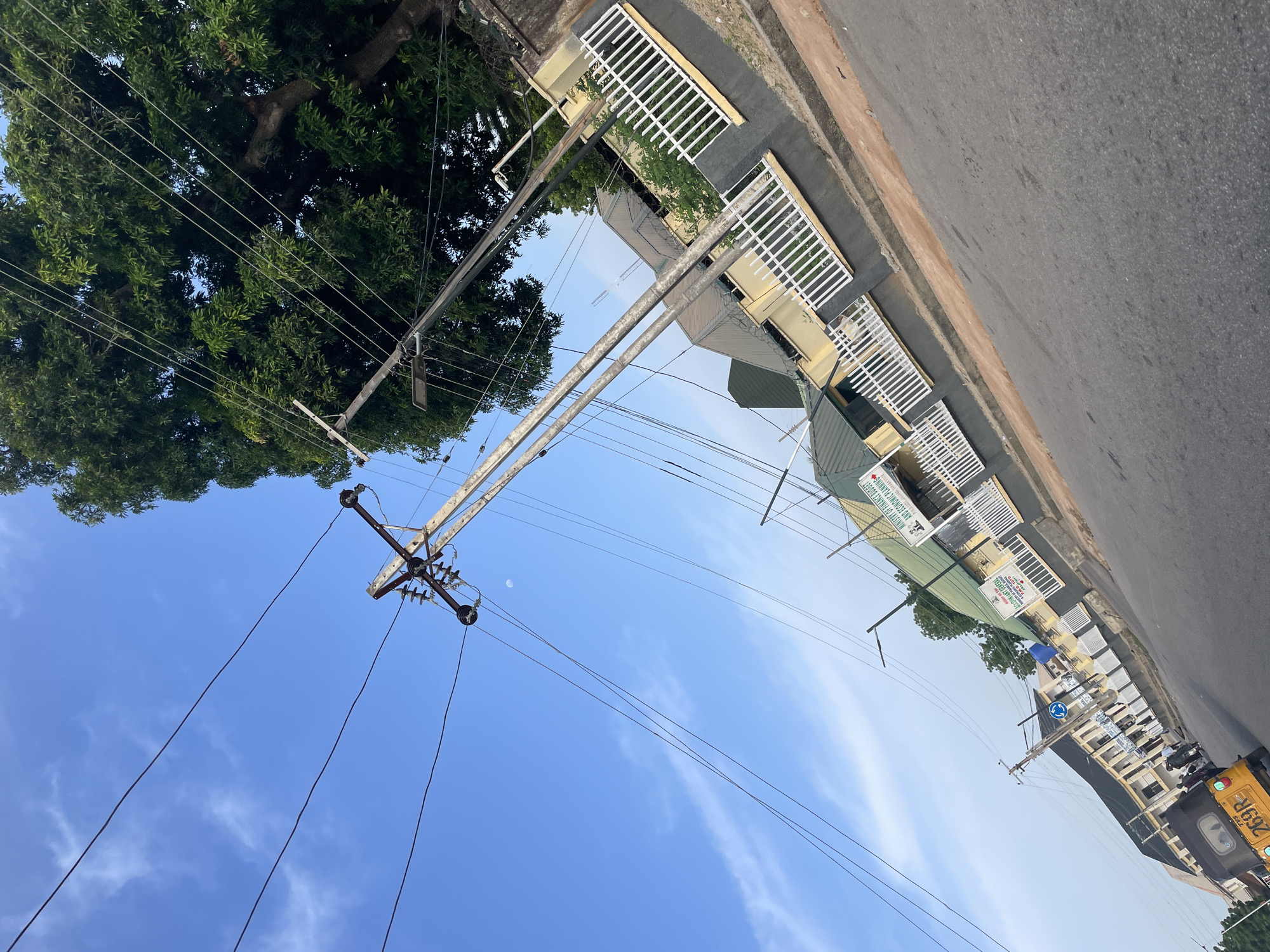 The image shows a street scene in Jalingo, Nigeria, featuring overhead power lines and a large tree on the right. A building with a white fence in front of it is visible behind the power lines. The building has signs, one of which reads "MINISTRY OF FINANCE BUDGET AND ECONOMIC PLANNING." A yellow motorized tricycle, commonly known as an 'okada' or 'keke napep', with the license plate "269R" is parked on the left side of the street, near a group of people. The sky is blue with some clouds. The image is taken at an angle, giving a tilted perspective of the scene.