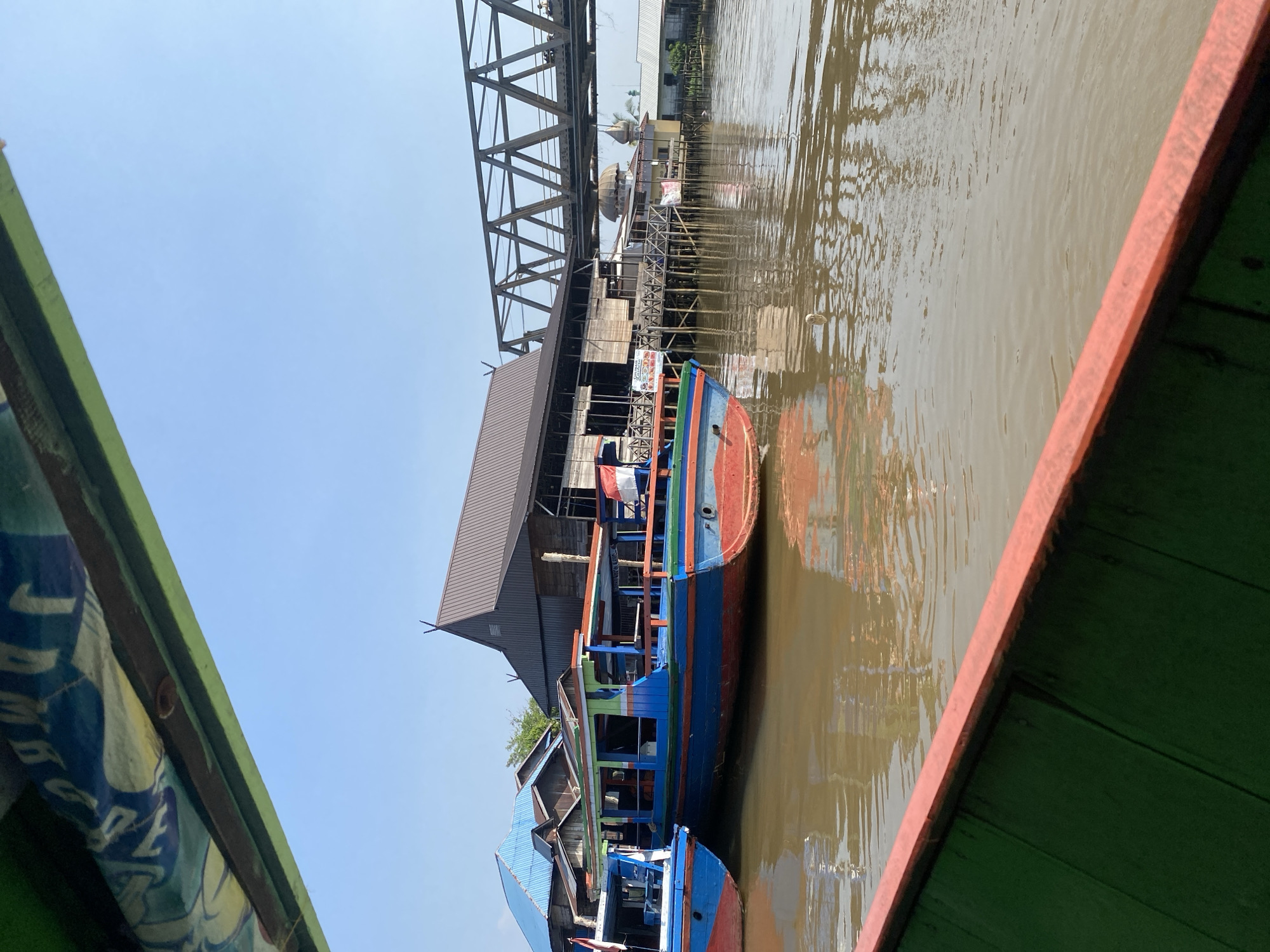 This image captures a scene from the perspective of someone on a boat in the city of Banjarmasin, Indonesia. The foreground shows the green and red railing of the boat. In the middle ground, two colorful wooden boats, predominantly blue and red with green accents, are moored side-by-side in murky brown water. Behind the boats, several wooden buildings with pitched roofs are visible, some with blue metal roofing. Further back, a large metal truss bridge spans across the water. The sky is a clear, bright blue. The scene depicts a typical waterway and settlement in Banjarmasin, a city known for its extensive network of rivers and canals.