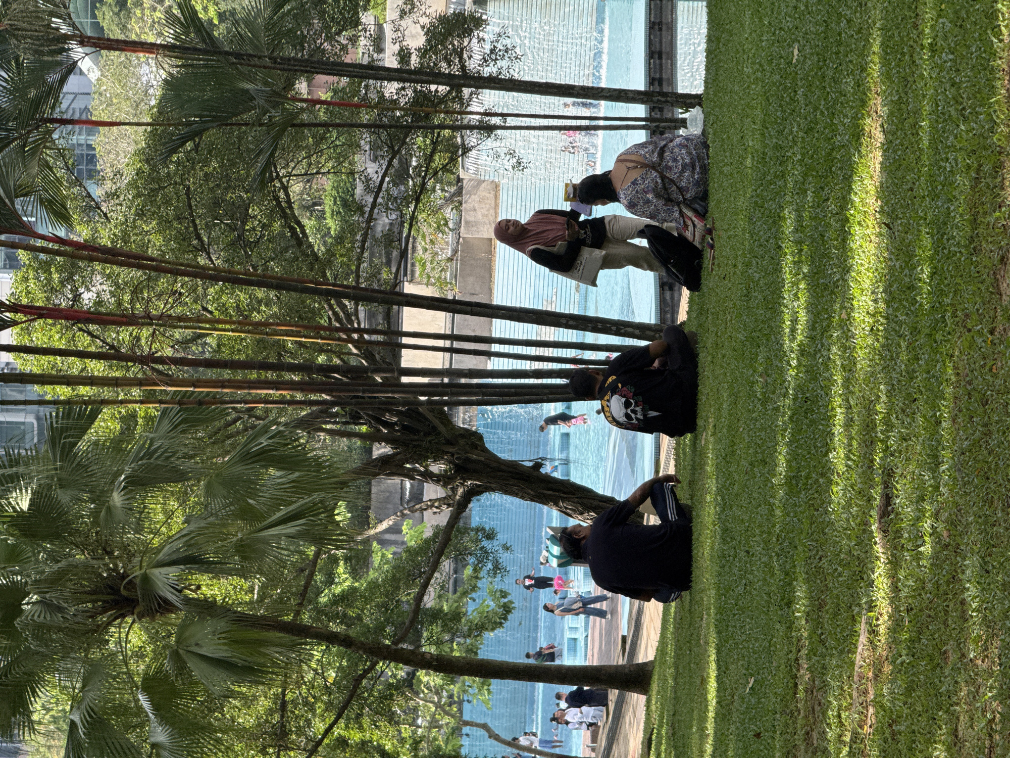 The image shows a public space in Kuala Lumpur, Malaysia, with a body of water, likely a decorative pool or fountain. Several people are present, some standing or sitting near the water's edge, others inside the water. Some appear to be interacting with each other, while others are solitary. The environment includes green grass, palm trees, and taller buildings partially visible in the background. People are dressed in casual clothing. Some women are wearing hijabs. Some people in the water appear to be playing.