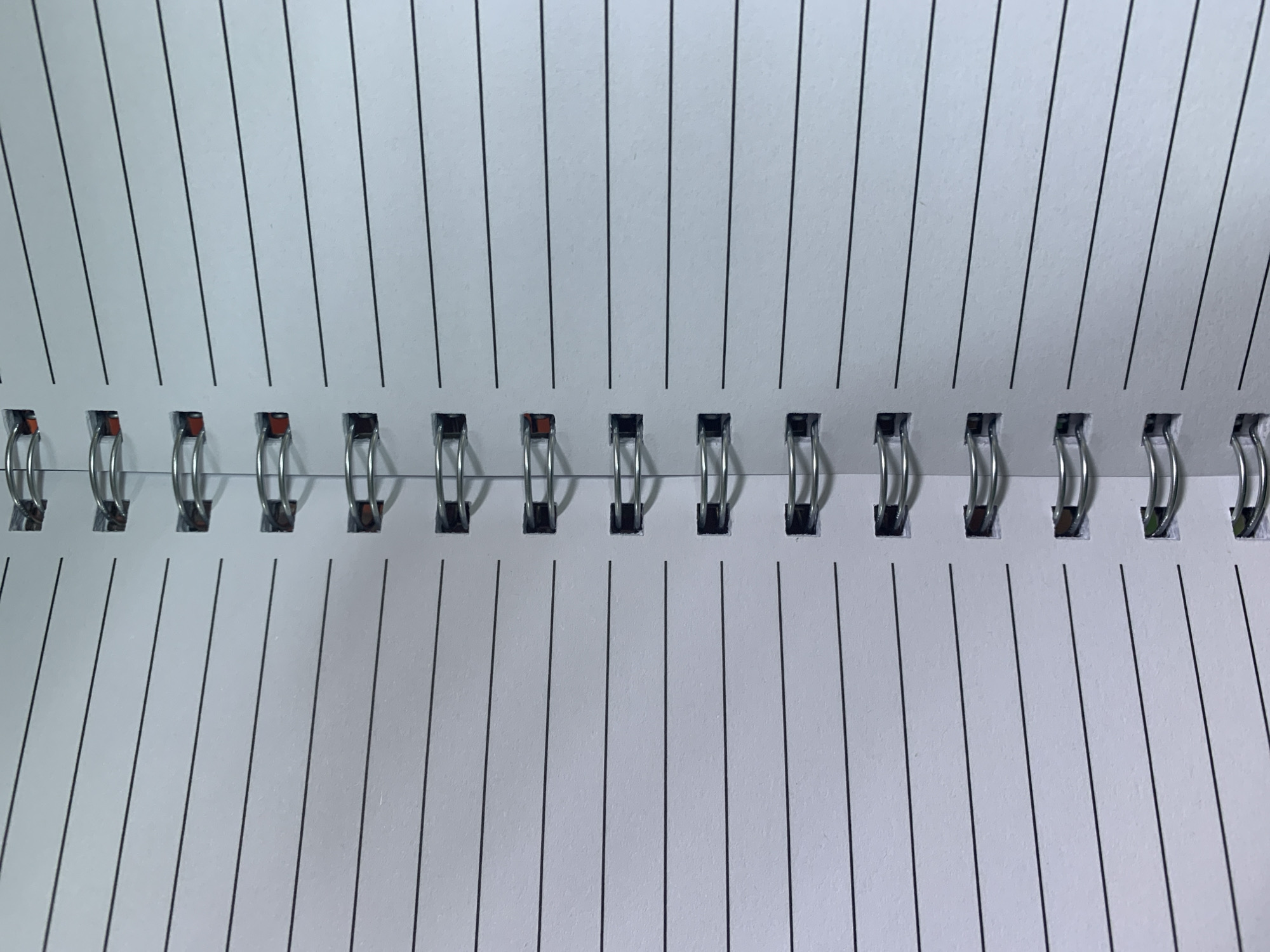 This is a close-up, high-angle, eye-level shot of a spiral-bound notebook. The notebook is open, revealing two pages with faint horizontal blue lines. The binding is a silver spiral that runs vertically down the center of the image. The notebook appears to be placed on a flat surface, though the surface itself is not visible. There are no people or other objects in the frame.