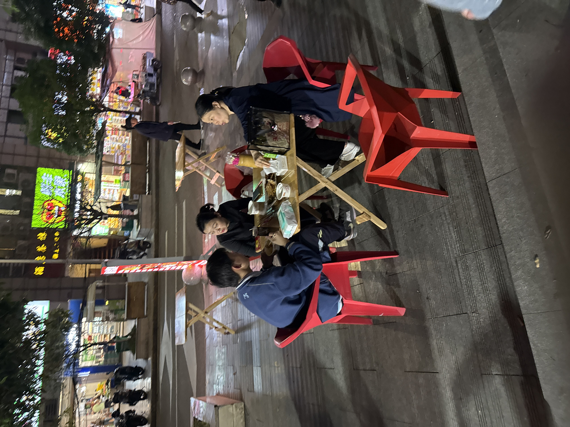 The image shows a street scene in the Shapingba District of Chongqing, China. Three people are seated at a small wooden table on a sidewalk. A woman in a dark jacket sits on a red plastic chair to the left, while a woman in a dark jacket sits across from her. A boy in a blue jacket is seated to the right. The table has food items on it. Red plastic chairs surround the table. In the background, there are buildings with lights and trees. The sidewalk appears to be wet. Pedestrians walk by in the background.
