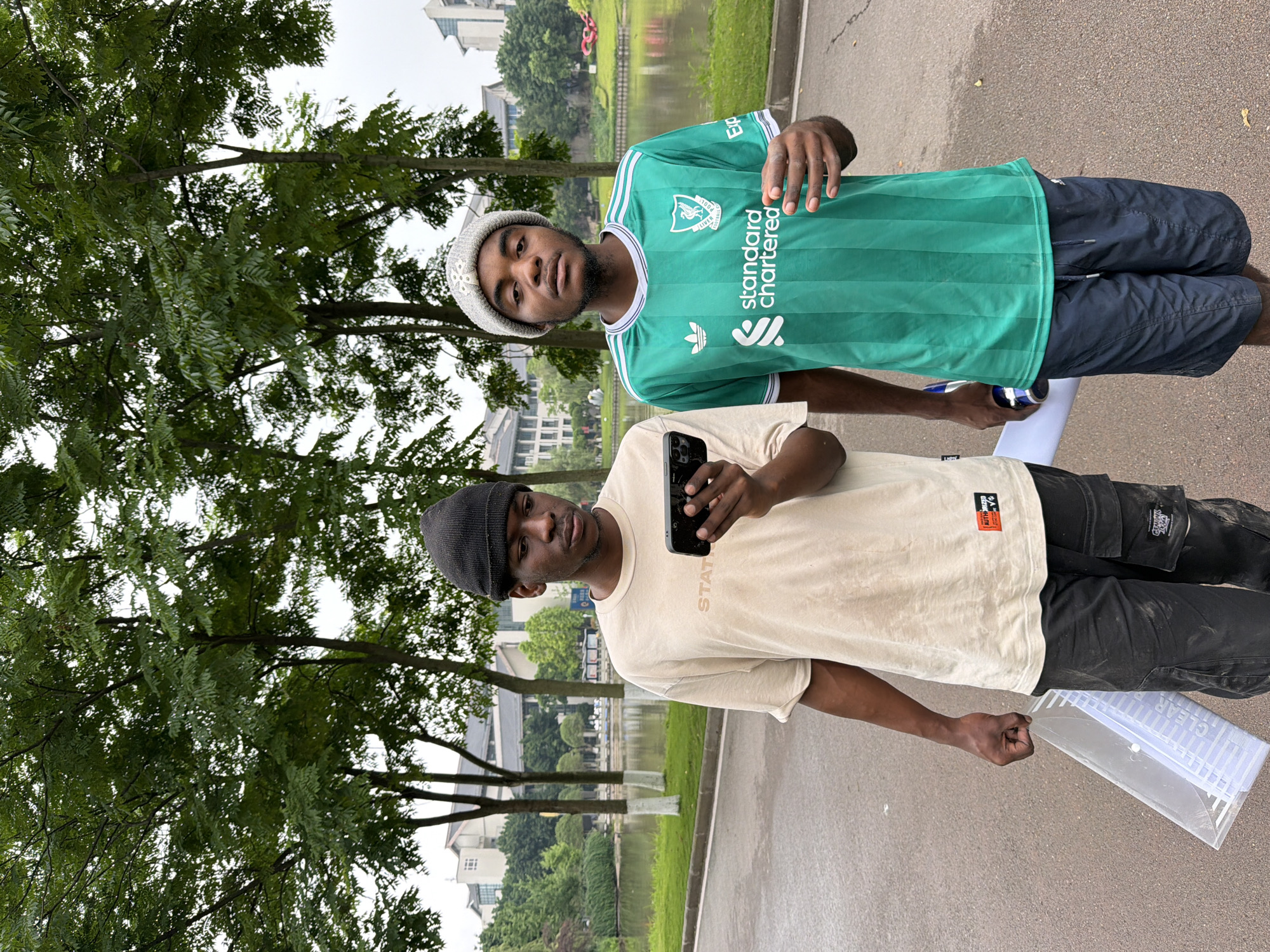 Two young Black men are standing on a paved walkway next to a body of water. The man on the left is wearing a beige t-shirt, black beanie, and black pants, and is holding a smartphone. The man on the right is wearing a green and white striped soccer jersey and dark blue shorts, and is holding a rolled-up piece of paper. Behind them are trees and buildings. The scene appears to be in a park or campus area in Jiangjin District, Chongqing, China.