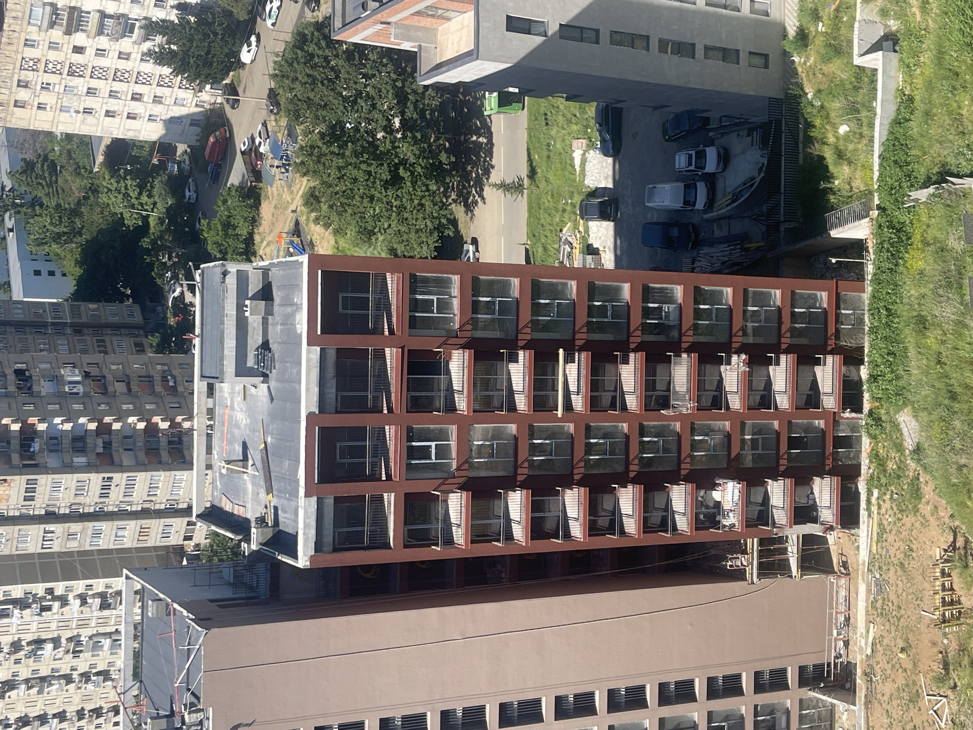 This is a medium-angle aerial shot of a city scene, likely Tbilisi, Georgia, depicting urban development and residential areas. The primary focus is on several multi-story buildings. A prominent feature is a modern, dark red building with numerous balconies and large windows, undergoing construction. To its left is a taller, pale brown building with uniformly spaced windows, also appearing to be residential. Further in the background, to the right and in the upper left, are older, light-colored apartment blocks with multiple floors and numerous small windows, exhibiting signs of wear.
In the foreground, the ground is a mix of construction debris, bare earth, and overgrown green grass. A narrow, paved road is visible in the middle ground, with several parked cars and a red minivan. A small playground with colorful structures is situated in a grassy area between the buildings. To the right of the red construction building, a lighter-colored building with a rooftop terrace is visible. A bright green dumpster is present near the road. The overall impression is one of an active urban environment with ongoing construction and established residential areas.