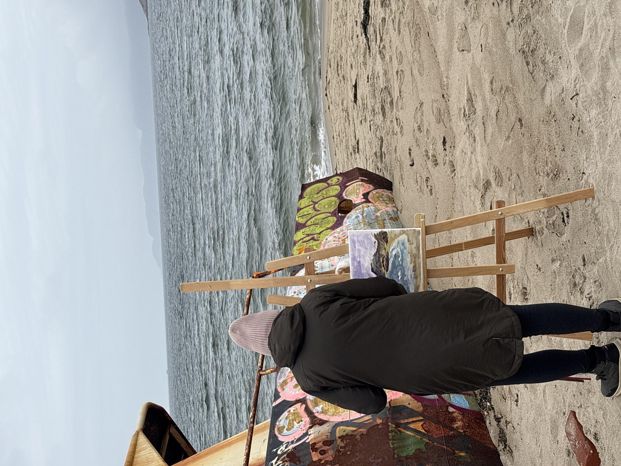 A person wearing a dark jacket and a light pink knitted hat stands on a sandy beach, facing away from the camera. They are positioned in front of an easel, on which a small painting is propped up. The painting appears to depict a landscape with water and hills. To the left of the person and easel is a large, rusty metal structure, possibly a shipwreck, covered in colorful graffiti. To the right, another section of graffiti-covered metal is visible, lying on the sand. The sea stretches out behind the beach, with waves gently rolling towards the shore. In the distance, a hazy coastline with hills can be seen under an overcast sky. The overall setting suggests a coastal location in Teriberka, Russia, known for its dramatic landscapes and shipwrecks.