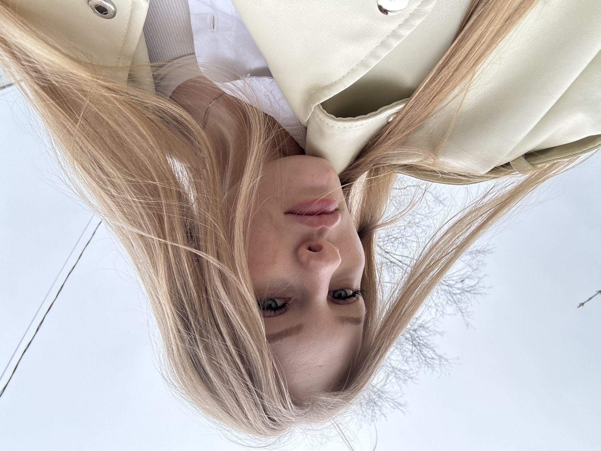 A young woman with blonde hair is lying down, looking directly at the camera. She has fair skin, green eyes, and is wearing a white shirt underneath a light green or beige jacket. Her hair is spread out around her head, and some strands are obscuring the background. The background is a pale sky with the bare branches of trees visible. This image seems to be taken outdoors in Perm, Russia, given the user's request to include that location.