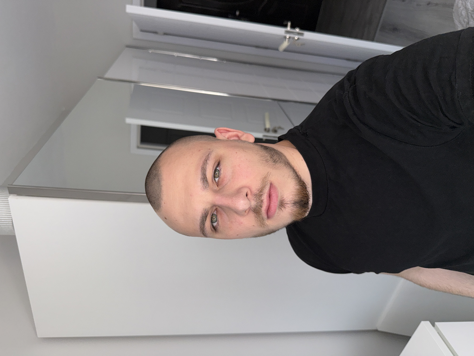 A headshot of a fair-skinned man with a shaved head, a beard, and green eyes is shown in a bathroom setting. He is wearing a black t-shirt. The background includes a white wall, a large mirror, a partially visible white cupboard with a silver handle, and a hint of gray flooring. No information regarding a location in Stara Zagora, Bulgaria, is available from the image.
