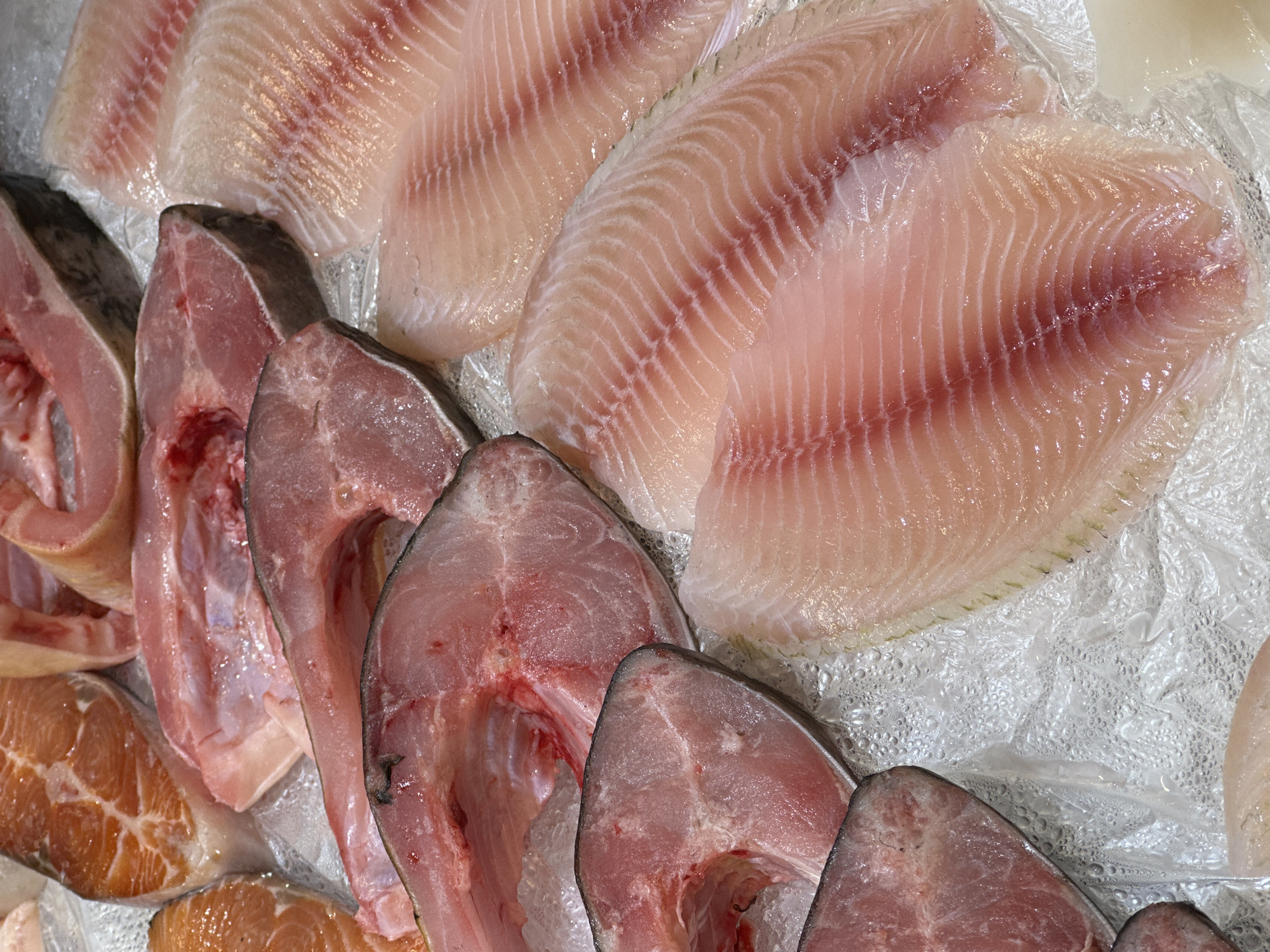 The image depicts a close-up view of raw fish fillets and steaks displayed on a bed of ice, likely at a market or fishmonger. The fillets are pale pink with distinct muscle striations, while the steaks are a darker red with visible bones and cut surfaces. The ice beneath is covered in small droplets of water. The location of the image is unknown, so the description will not be able to include any locations in the city of Khimki, Russia.