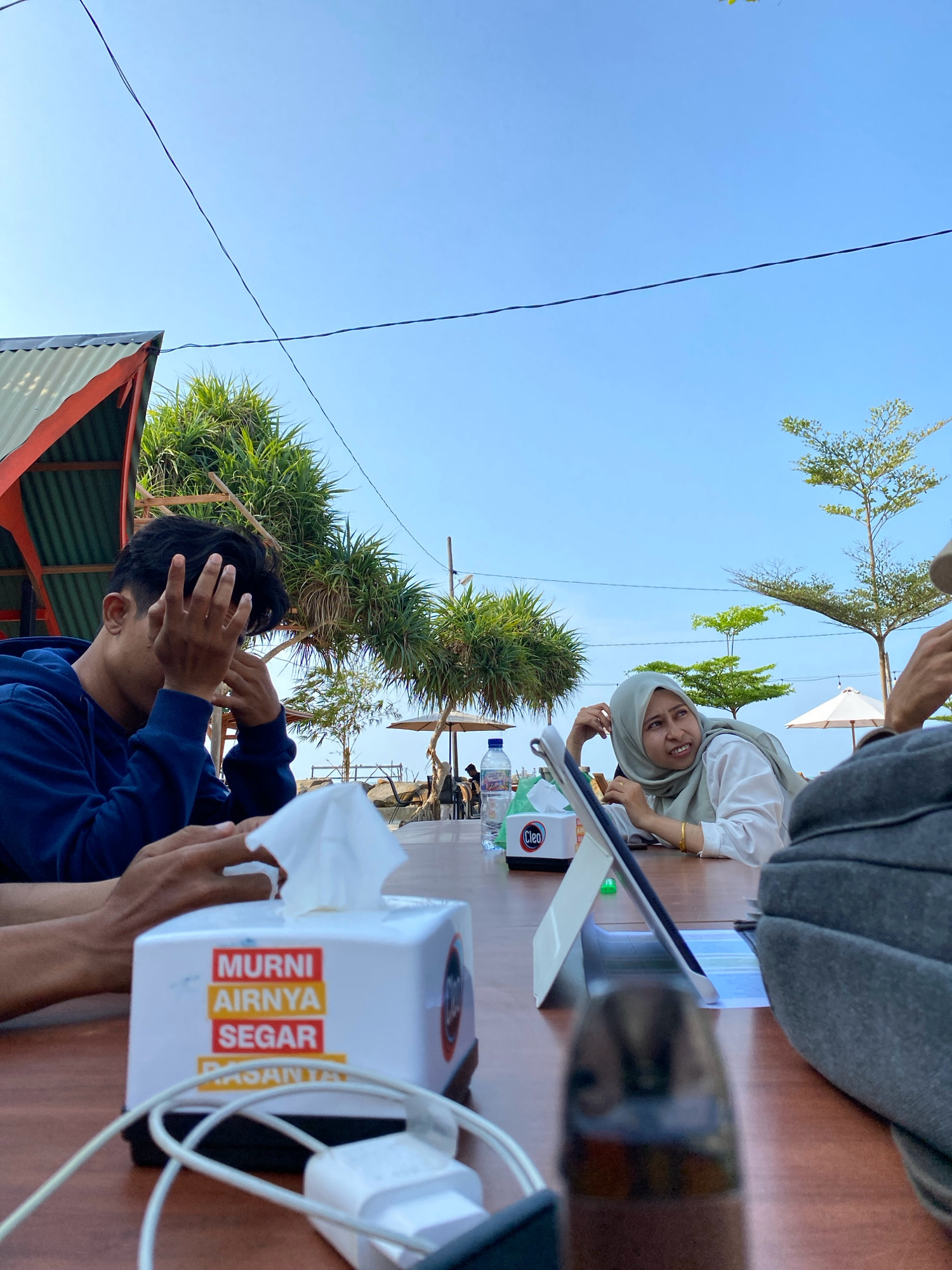 In Lancok, Indonesia, four individuals are seated at a wooden table outdoors, with a clear blue sky overhead and lush green trees framing the scene. A man in a blue hoodie is positioned on the left, with his hands near his face, appearing deep in thought or perhaps shielding his eyes. Across from him, a woman wearing a light grey hijab and a white shirt is looking to her right, with her right hand resting near her face. In the foreground, a tissue box with Indonesian text that translates to "Pure Fresh Water" is prominently displayed, alongside charging cables and a tablet. A grey bag is visible on the right side of the frame. The setting suggests a casual gathering or meal, possibly at a beachfront restaurant or cafe, given the palm-like trees and the distant hint of water.