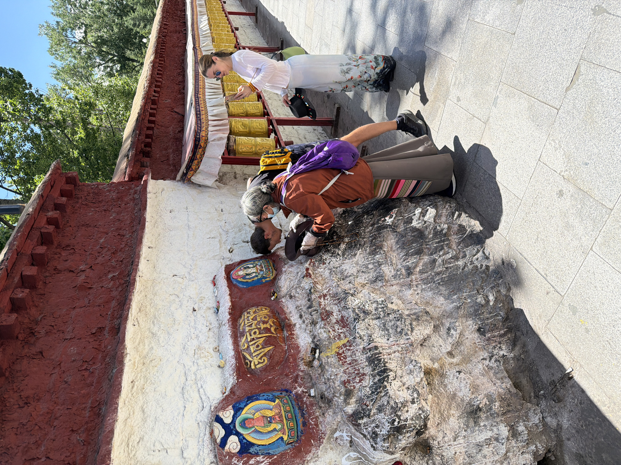 The image captures an outdoor scene on a bright, sunny day, likely at a Tibetan Buddhist site. Two individuals are present on a light grey tiled walkway next to a building or wall adorned with religious elements.
One person, appearing older with grey hair, is seen from behind, leaning forward intently towards a painted section of the wall. They are wearing a brown jacket, grey trousers with a colorful striped design at the hem, a purple backpack, and a face mask. Their hands are positioned near the base of the wall, where there are three colorful, painted or carved images. The central image clearly displays the Tibetan mantra "Om Mani Padme Hum" in golden script on a red background, flanked by two other painted figures resembling Buddhas or Bodhisattvas. The person's posture suggests a moment of contemplation, prayer, or veneration.
Standing slightly behind and to the right is another individual, dressed in a long-sleeved white top and a long white skirt with an elaborate floral and bird pattern at the bottom. They are wearing sunglasses and are positioned near a row of golden prayer wheels that line the wall. Their left hand appears to be resting near the prayer wheels, while they hold a black hat in their right hand.
The scene is illuminated by strong sunlight, casting distinct shadows on the ground. In the background, vibrant green trees are visible against a clear blue sky, indicating an outdoor, natural setting. The overall atmosphere suggests a moment of cultural or religious observance.